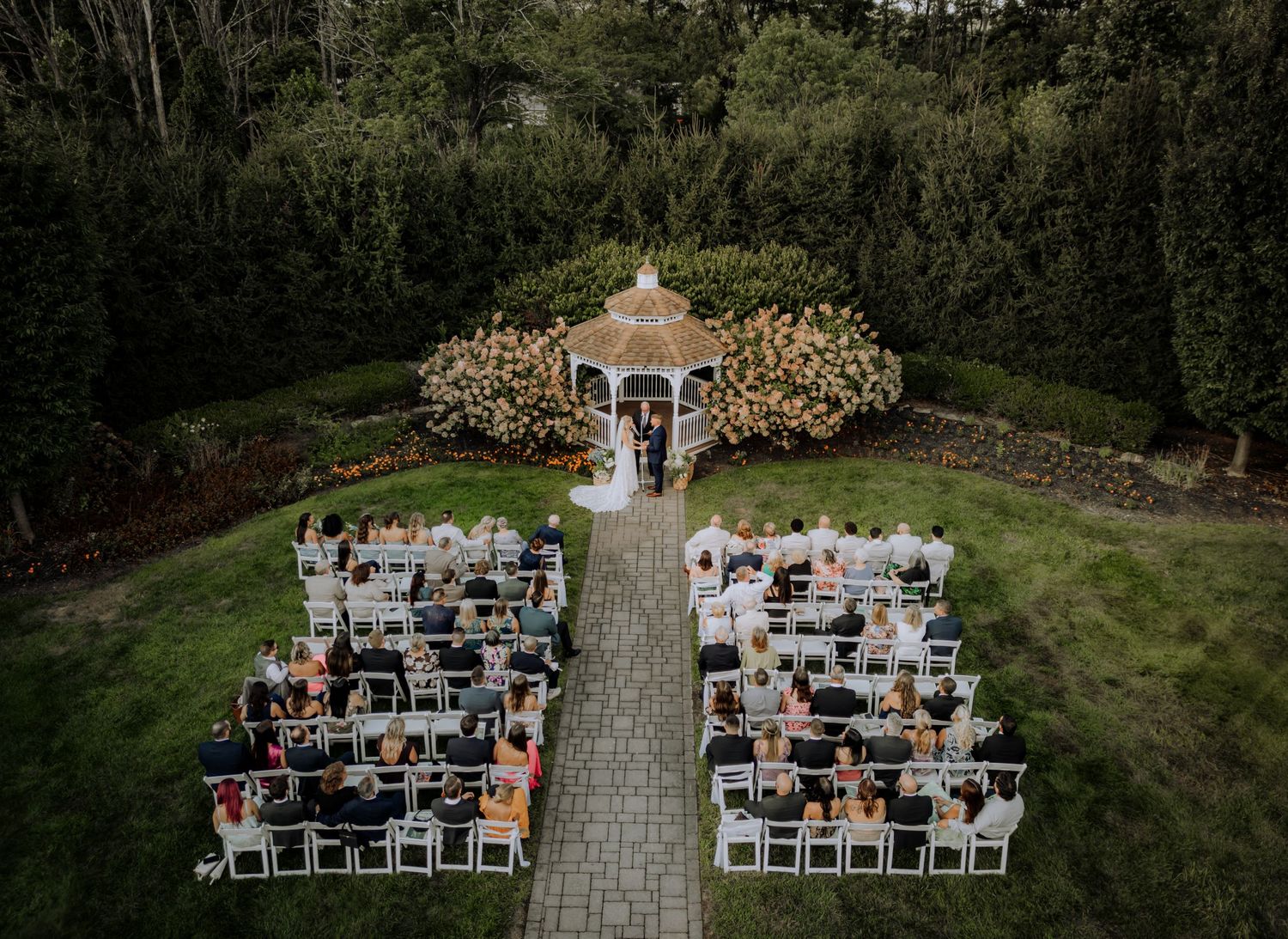 Aerial view of outdoor wedding ceremony with white chairs set up along stone pathway surrounded by lush greenery.