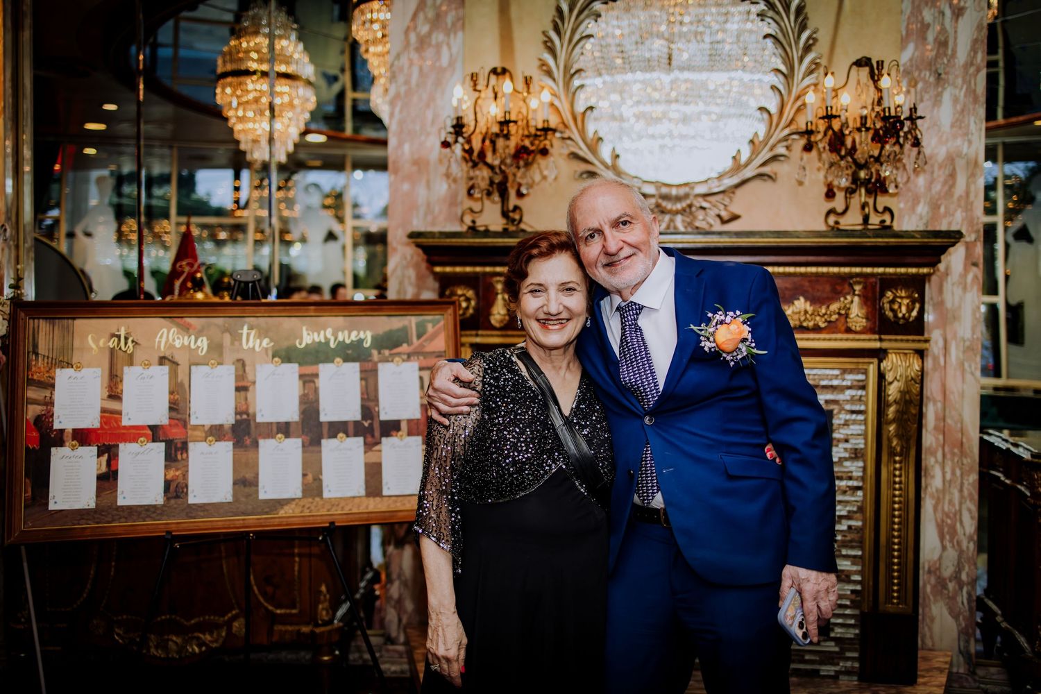 Wedding guests socialize in an ornately decorated reception hall with gold accents and crystal chandeliers.