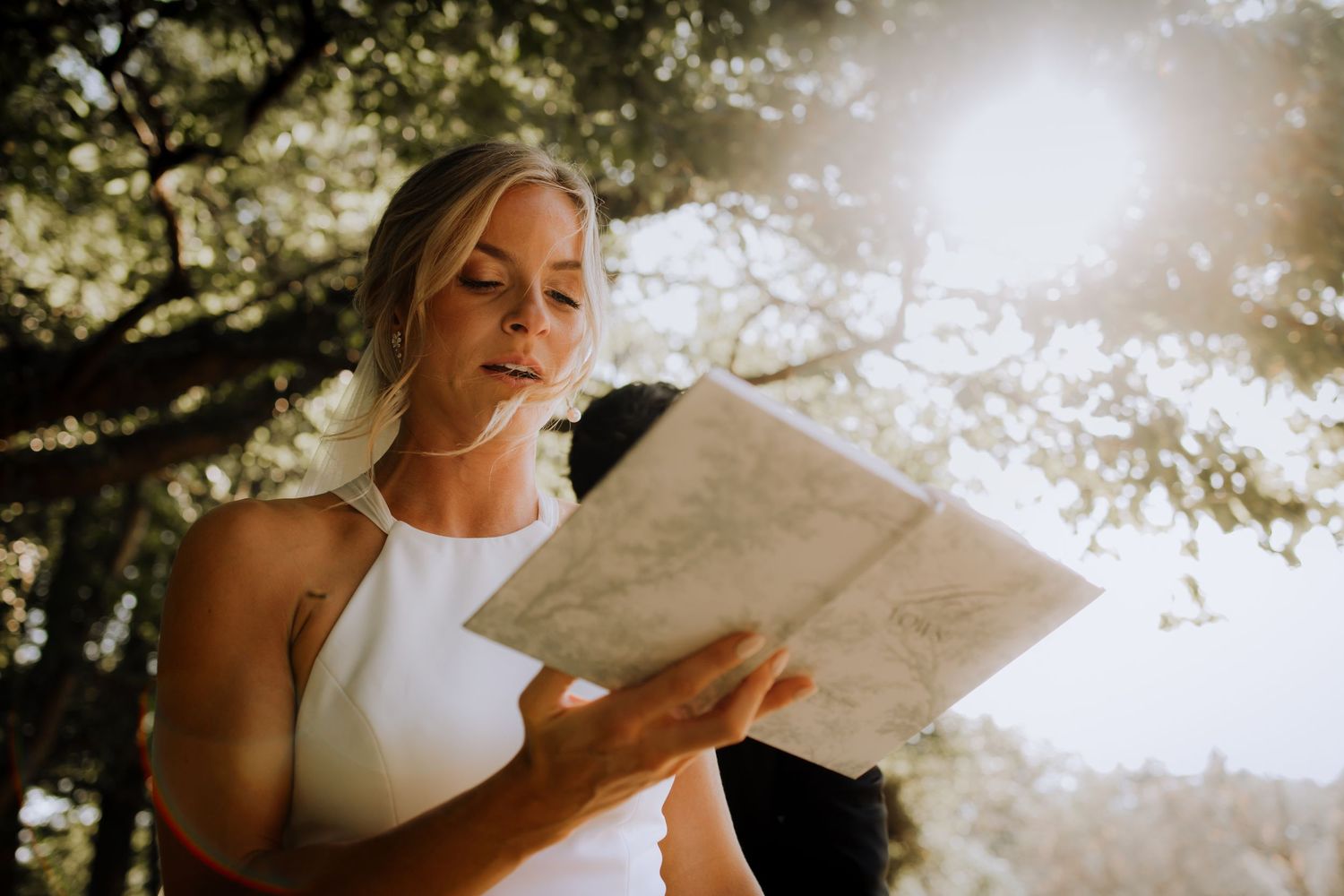 A bride in a white dress reads a letter outdoors with sunlight streaming through trees in the background.