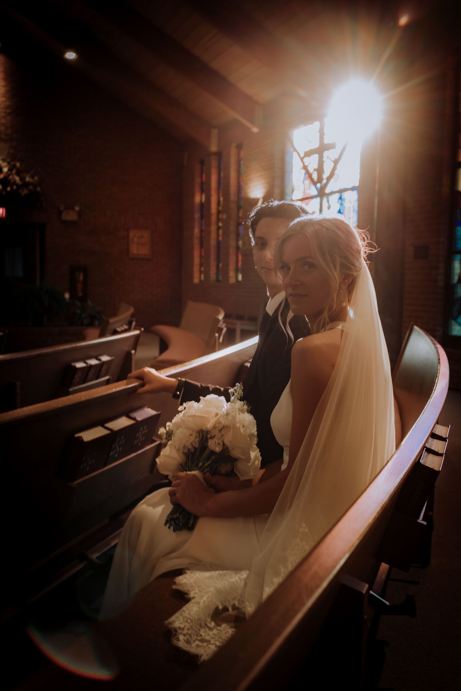 A couple sits together in a wooden church pew, with sunlight streaming through stained glass windows in warm, moody lighting.