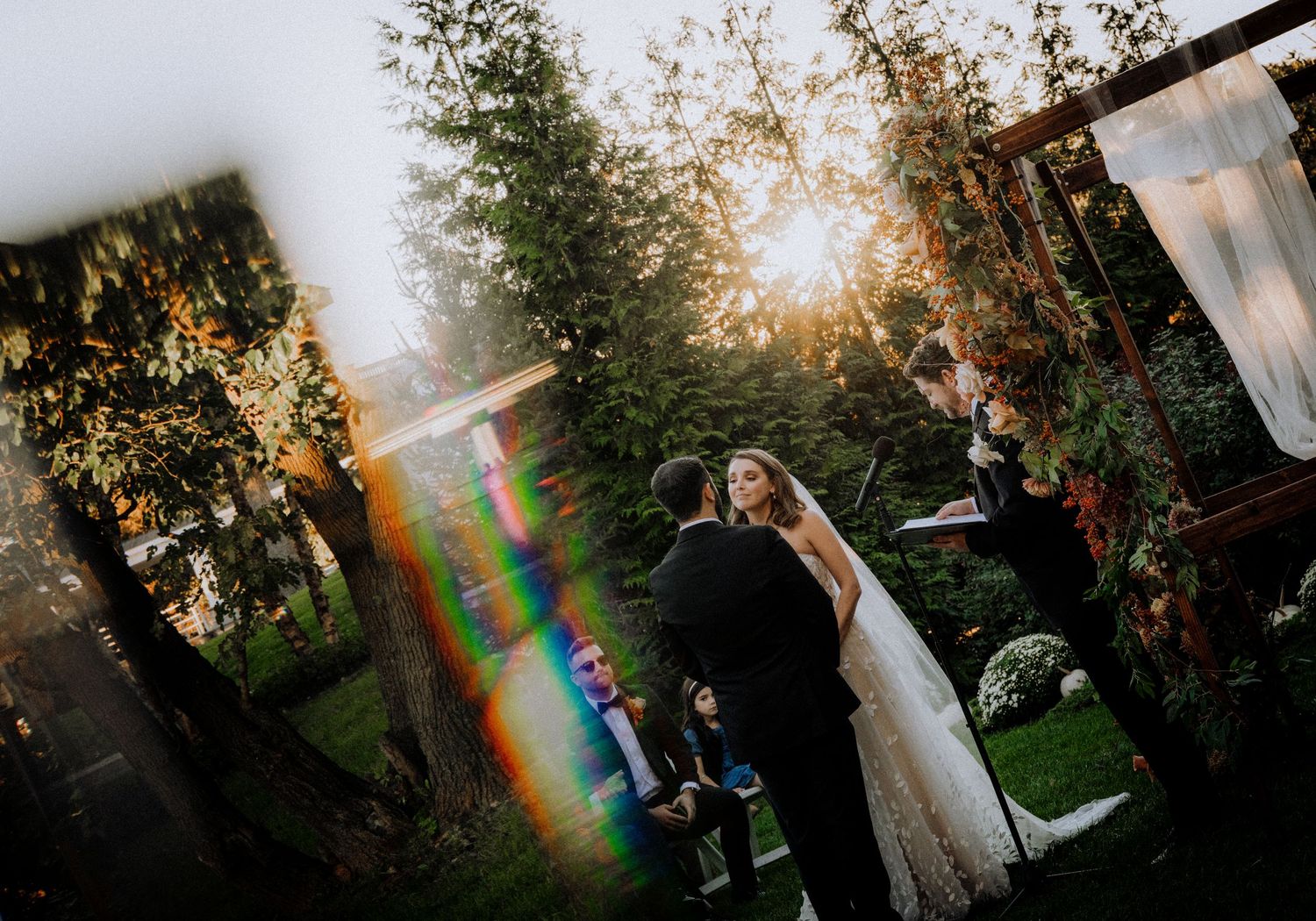 Outdoor wedding ceremony with dramatic sunset lens flare through trees.