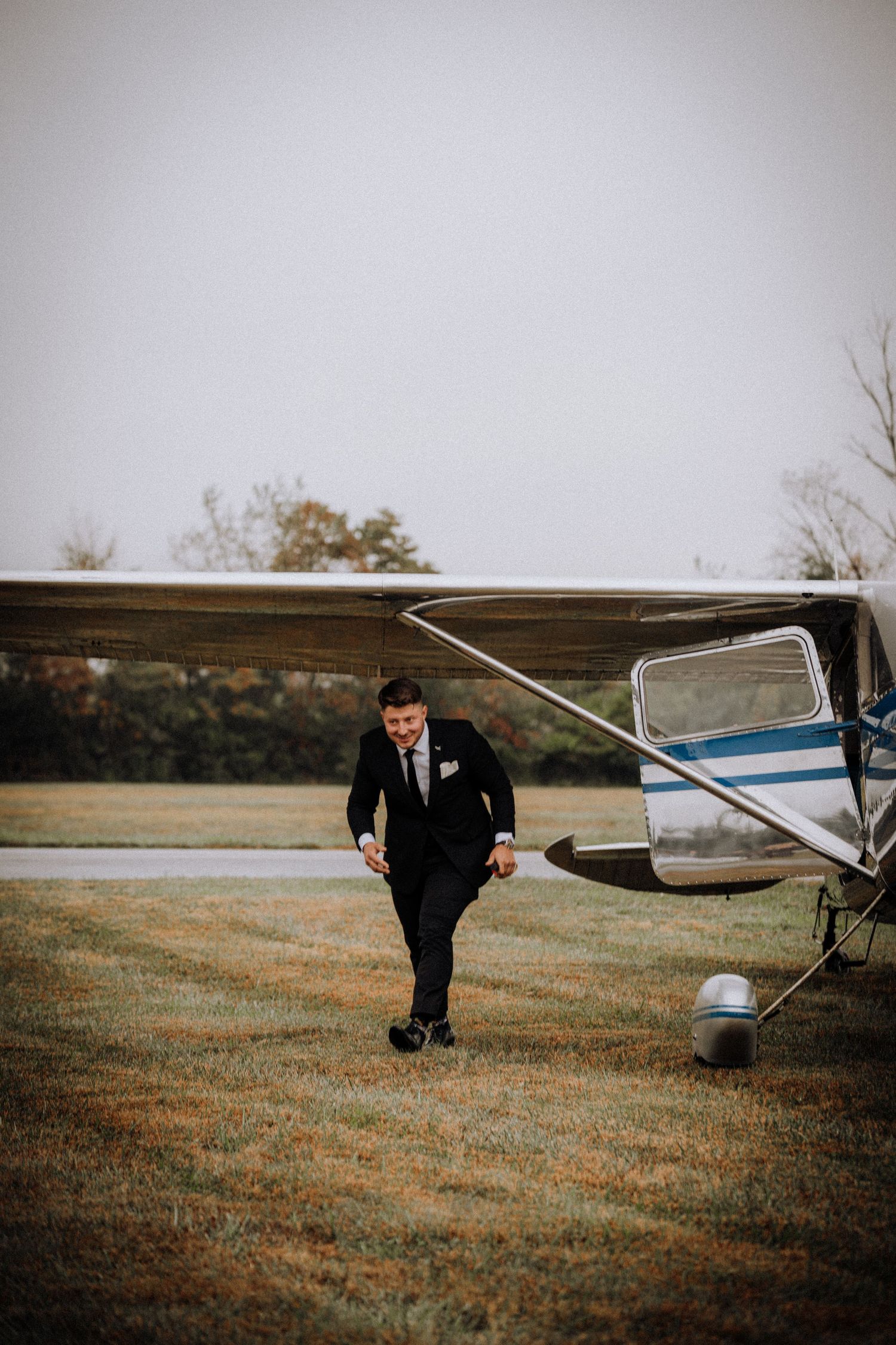 Wedding party member in a suit crouches near a parked airplane on a grassy field.
