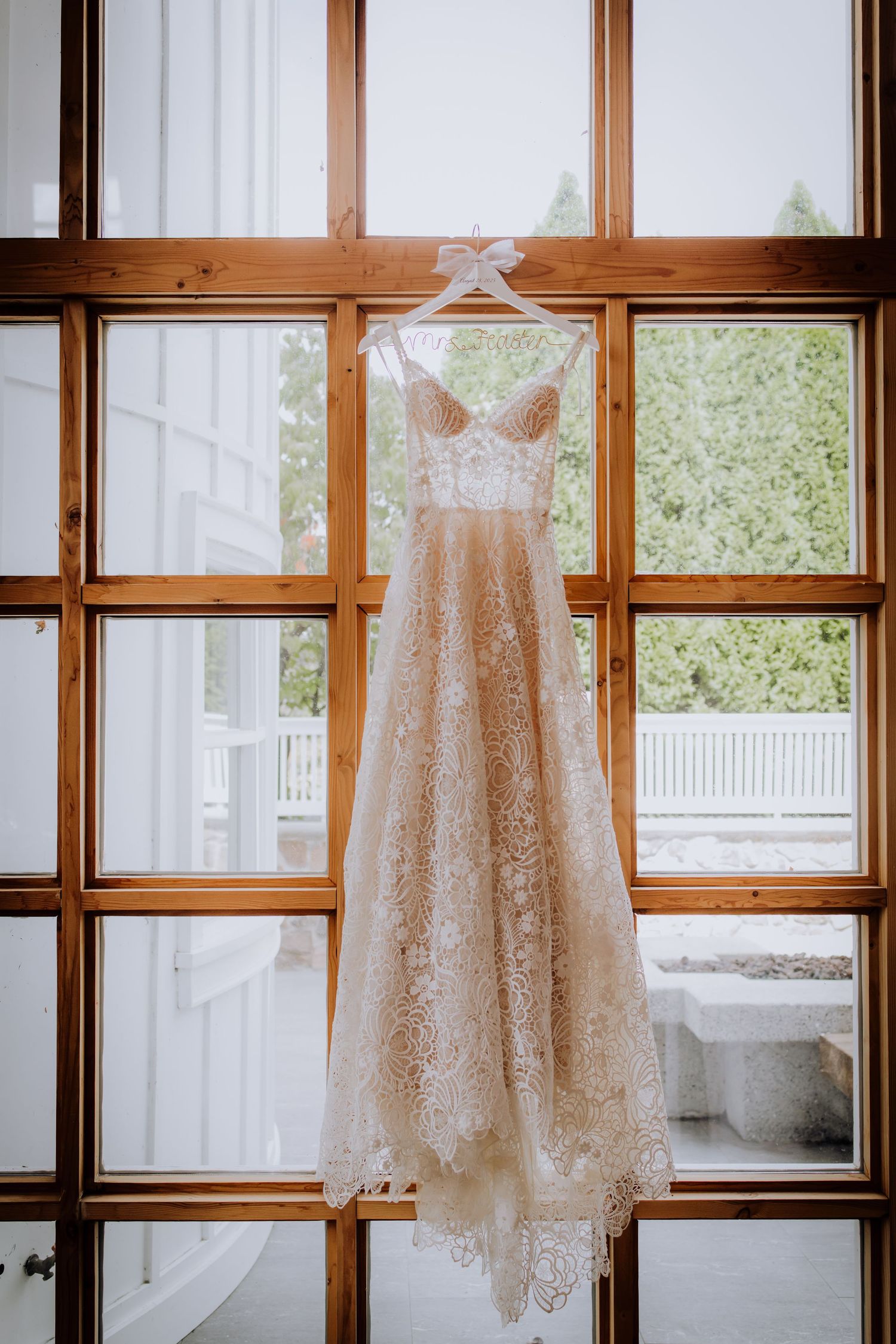 White lace wedding dress and accessories hanging against wooden window frames with natural light streaming in.