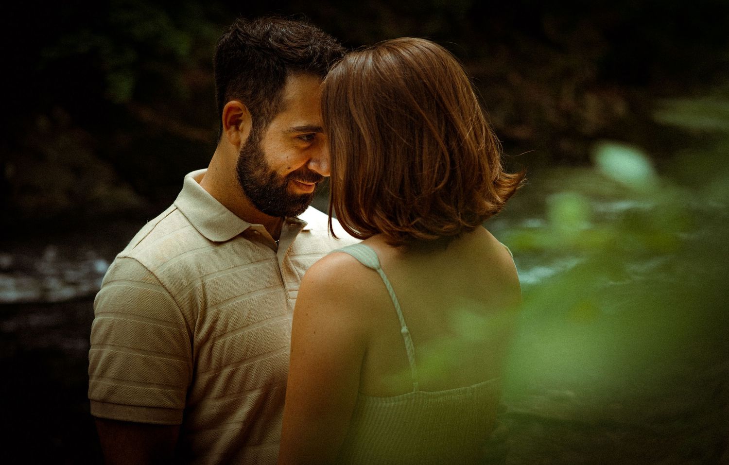 Intimate romantic couple sharing a tender moment in nature with soft bokeh lighting and green foliage in foreground.