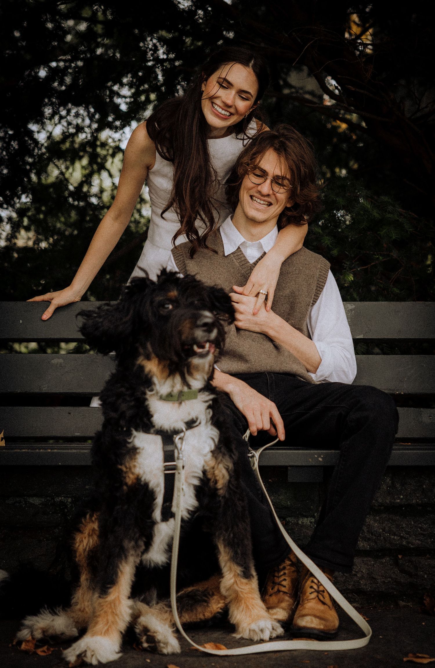 A happy couple sits on a bench with their large fluffy dog during an outdoor photo session.