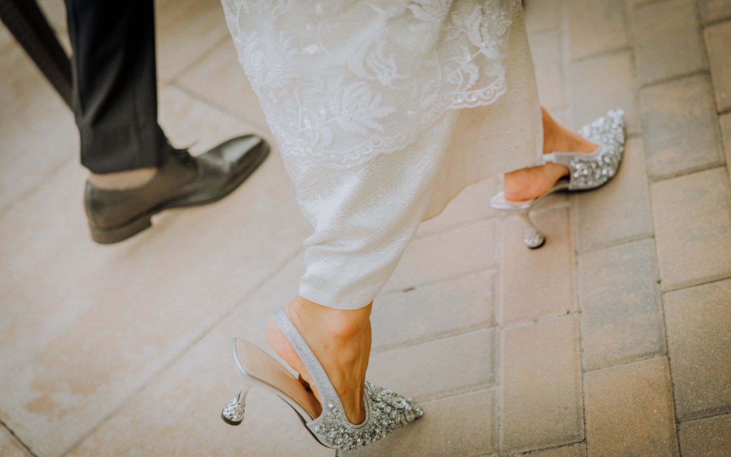 Close-up view of elegant sparkly silver bridal heels and groom's dress shoes on a light textured floor.
