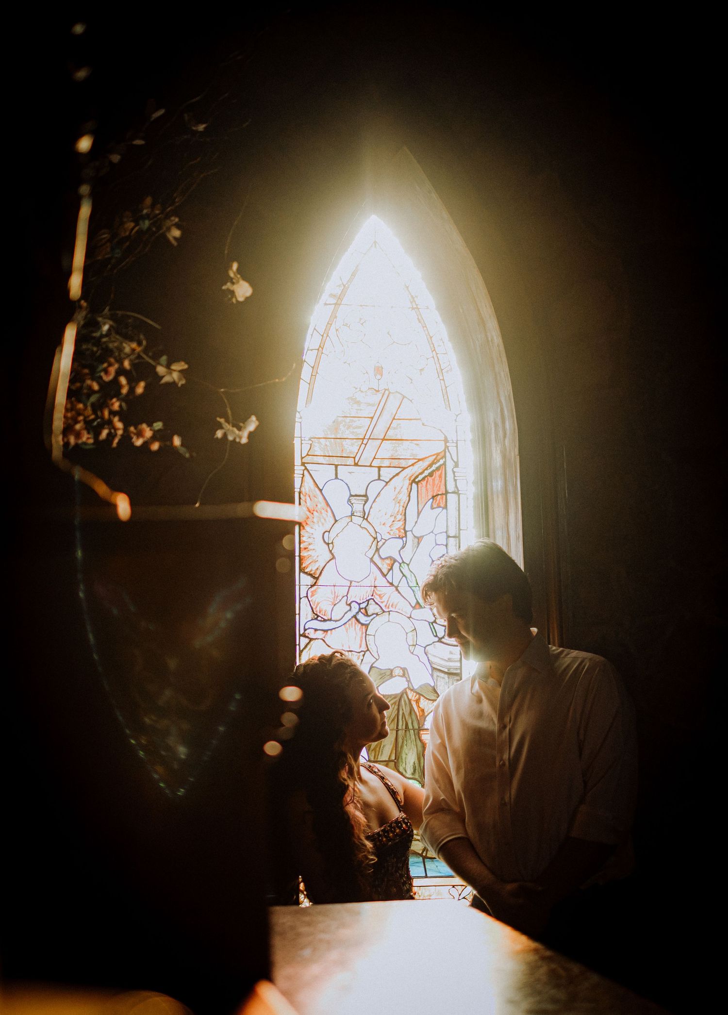 A silhouetted couple stands before a glowing stained glass church window.