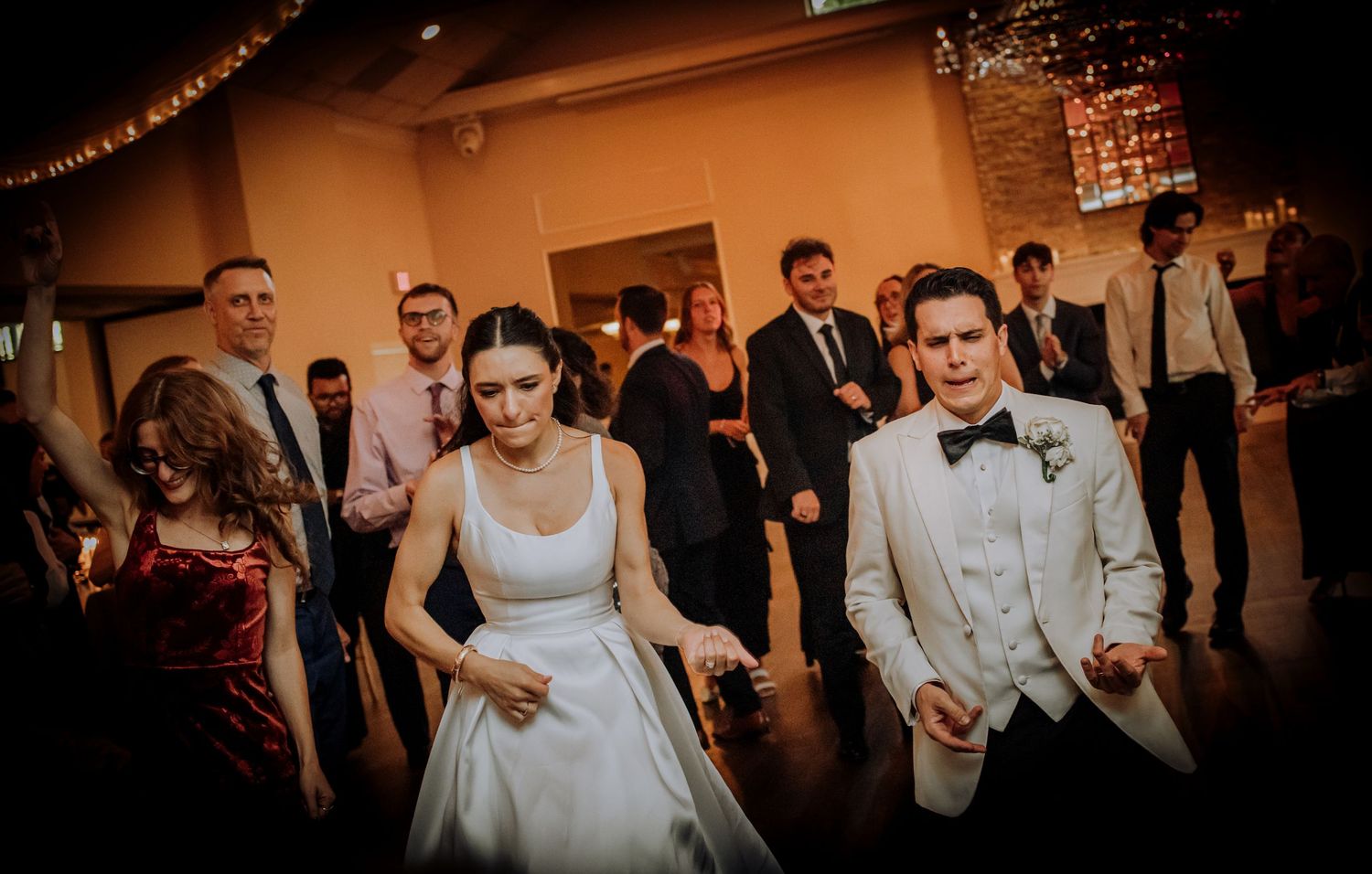 Wedding reception guests dancing energetically on a dimly lit dance floor decorated with string lights.