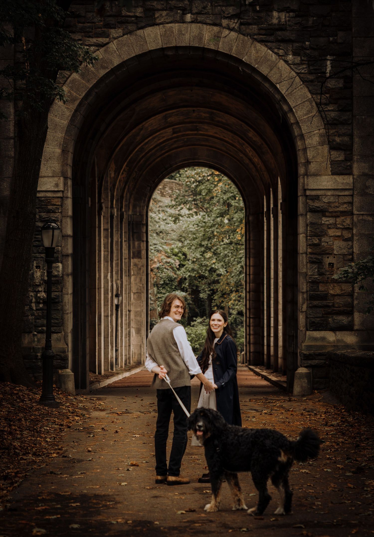 Couple strolls through a dramatic stone archway tunnel with natural light streaming in from the end.