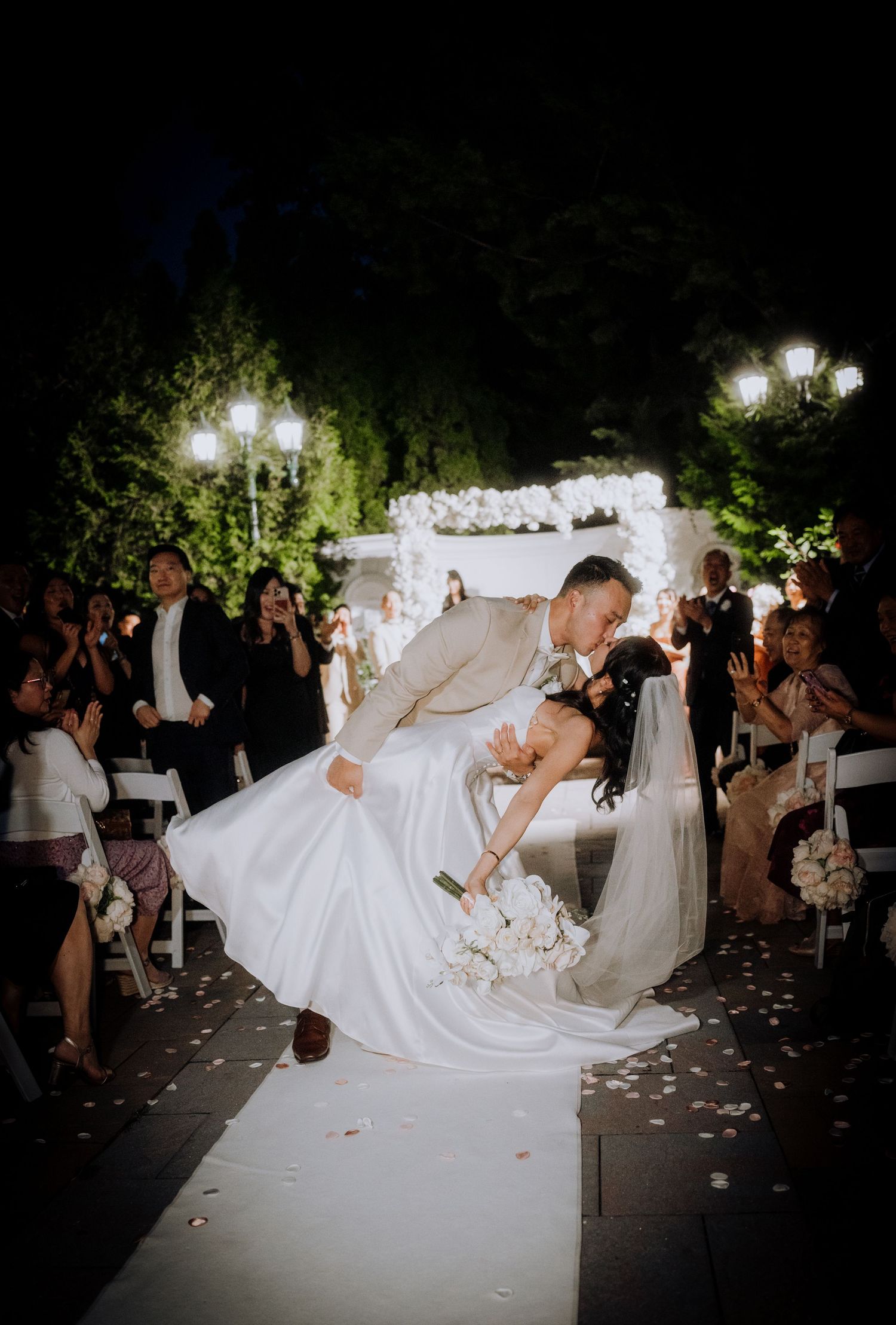 Dramatic wedding dip pose under evening lights with guests watching and flower petals scattered.