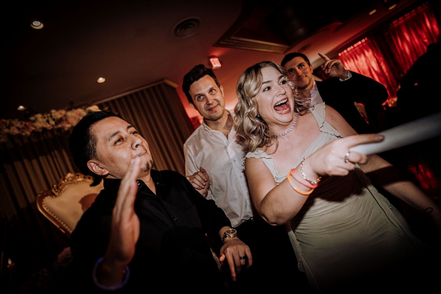 Wedding party guests dance and celebrate with joyful expressions at a reception under red lighting.