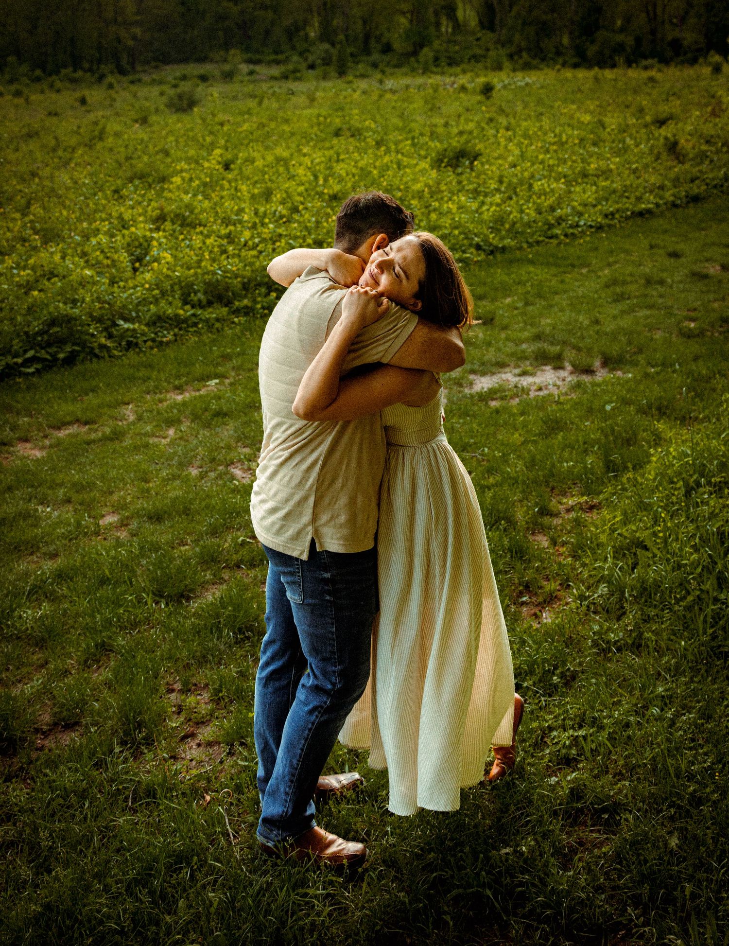 A couple embraces affectionately in a sunlit meadow with lush green grass and wildflowers in the background.
