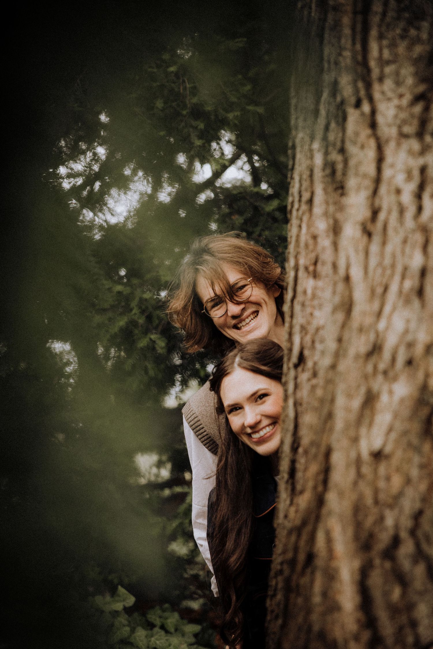 Two people peek playfully from behind a large tree trunk in a forest setting.
