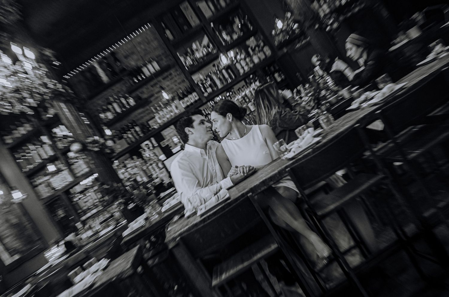 Blurred black and white view of a bar counter with bottles and glassware in moody lighting.