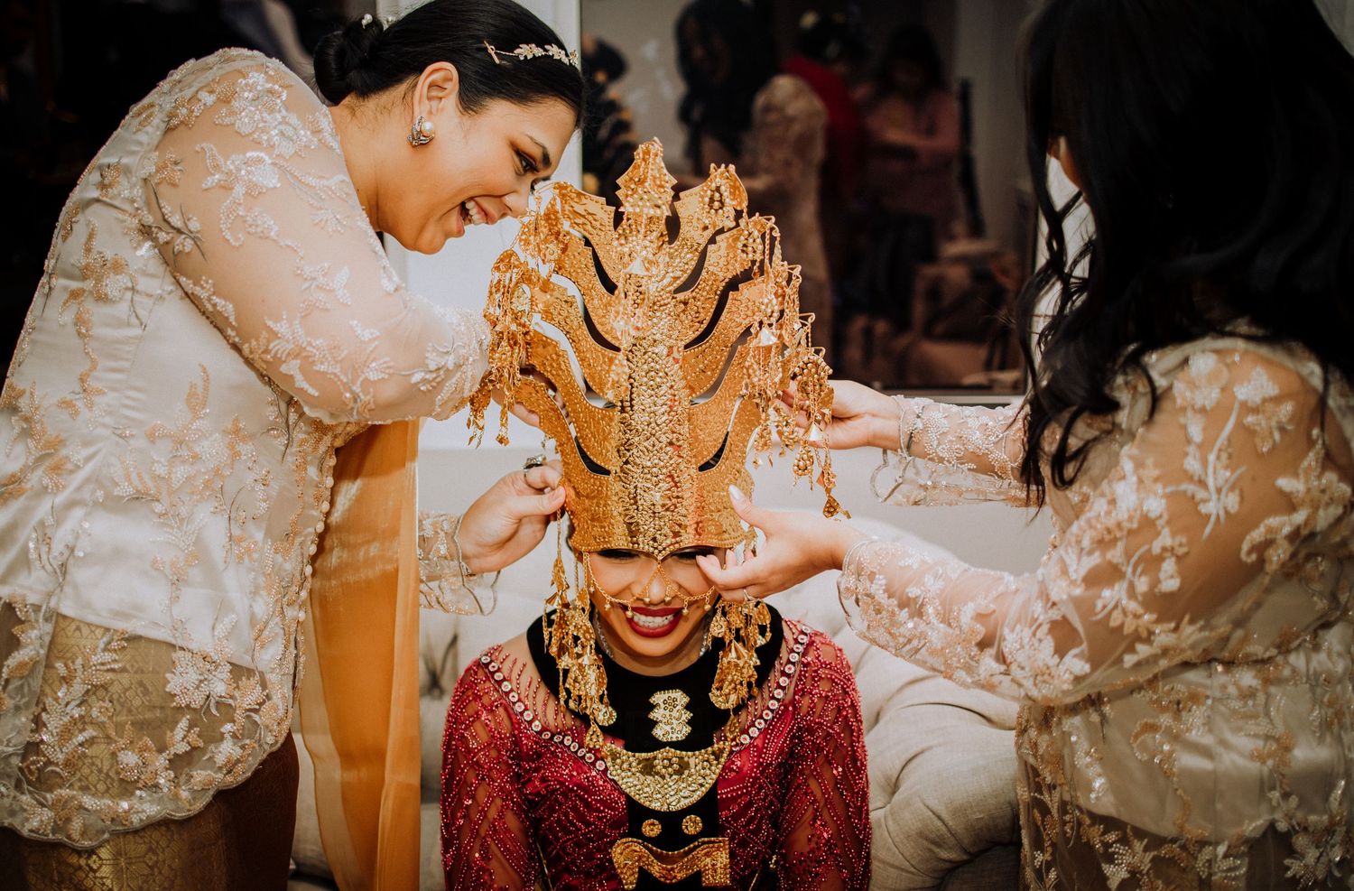 Traditional Indonesian ritual shows hands adorned with henna placing a golden crown headpiece on someone wearing red attire.