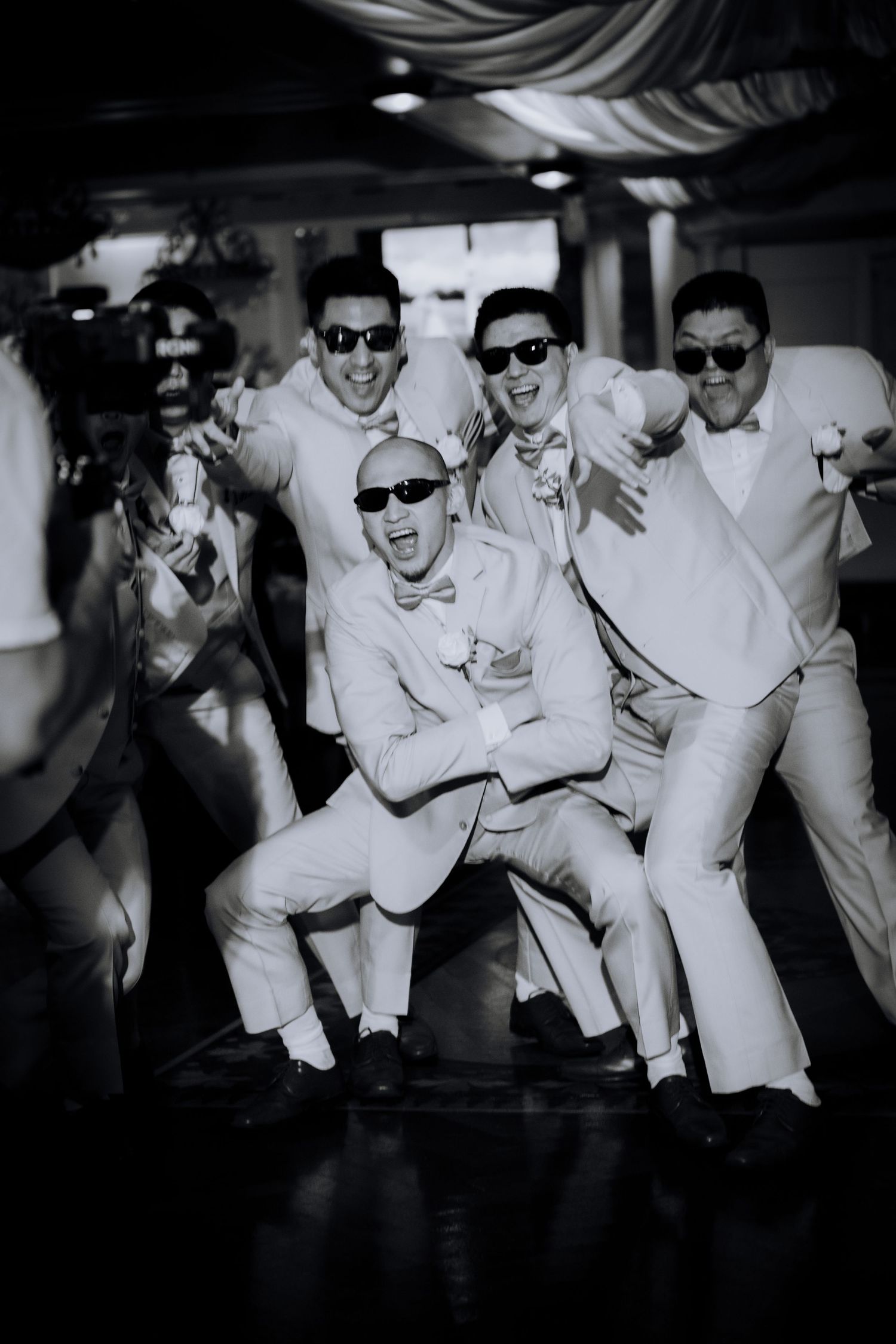 Groomsmen in white suits and sunglasses striking playful poses in black and white photography.