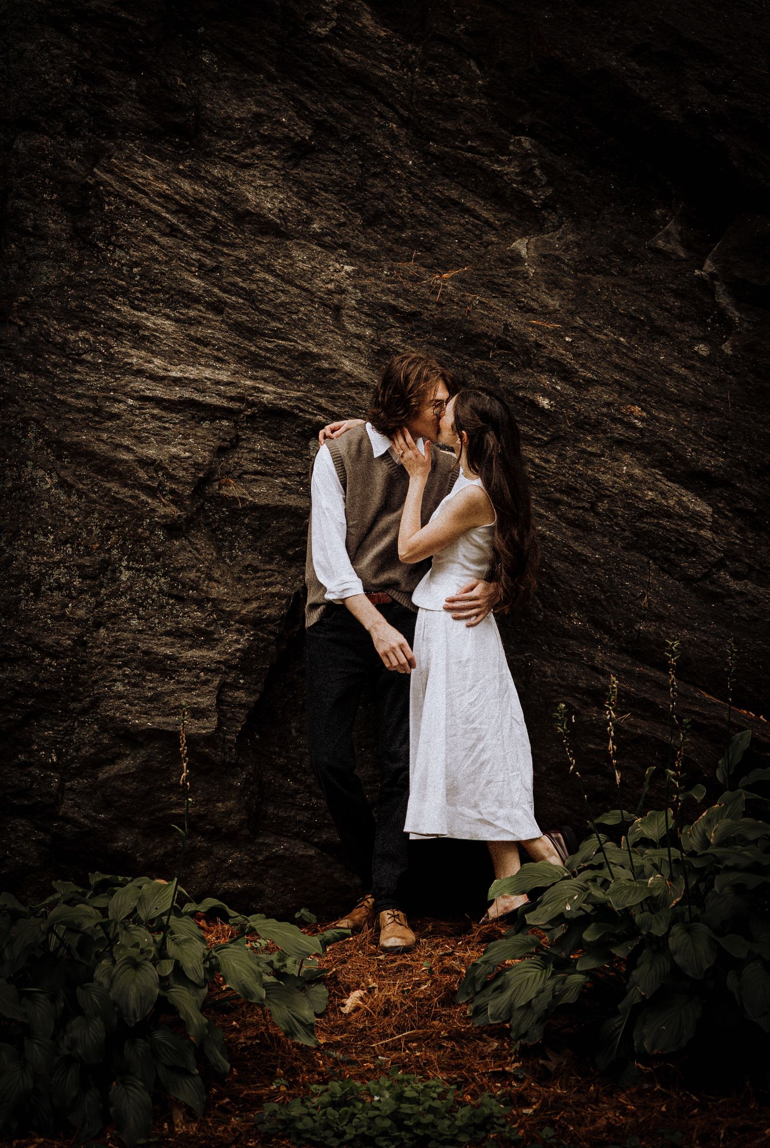Couple embraces intimately against dramatic dark rock wall with green foliage in moody outdoor setting.