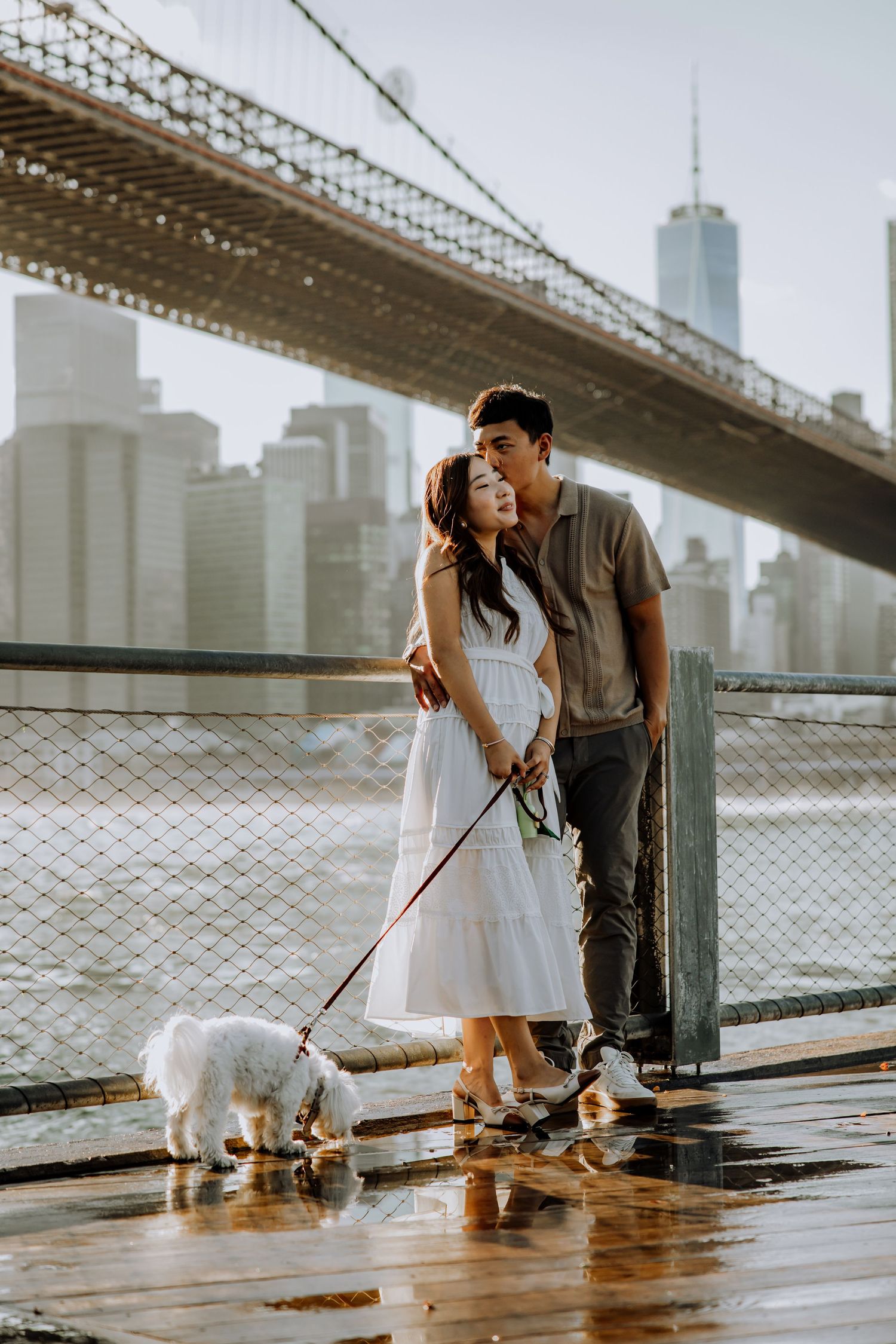 Couple with small dog enjoying moment under Brooklyn Bridge with NYC skyline.