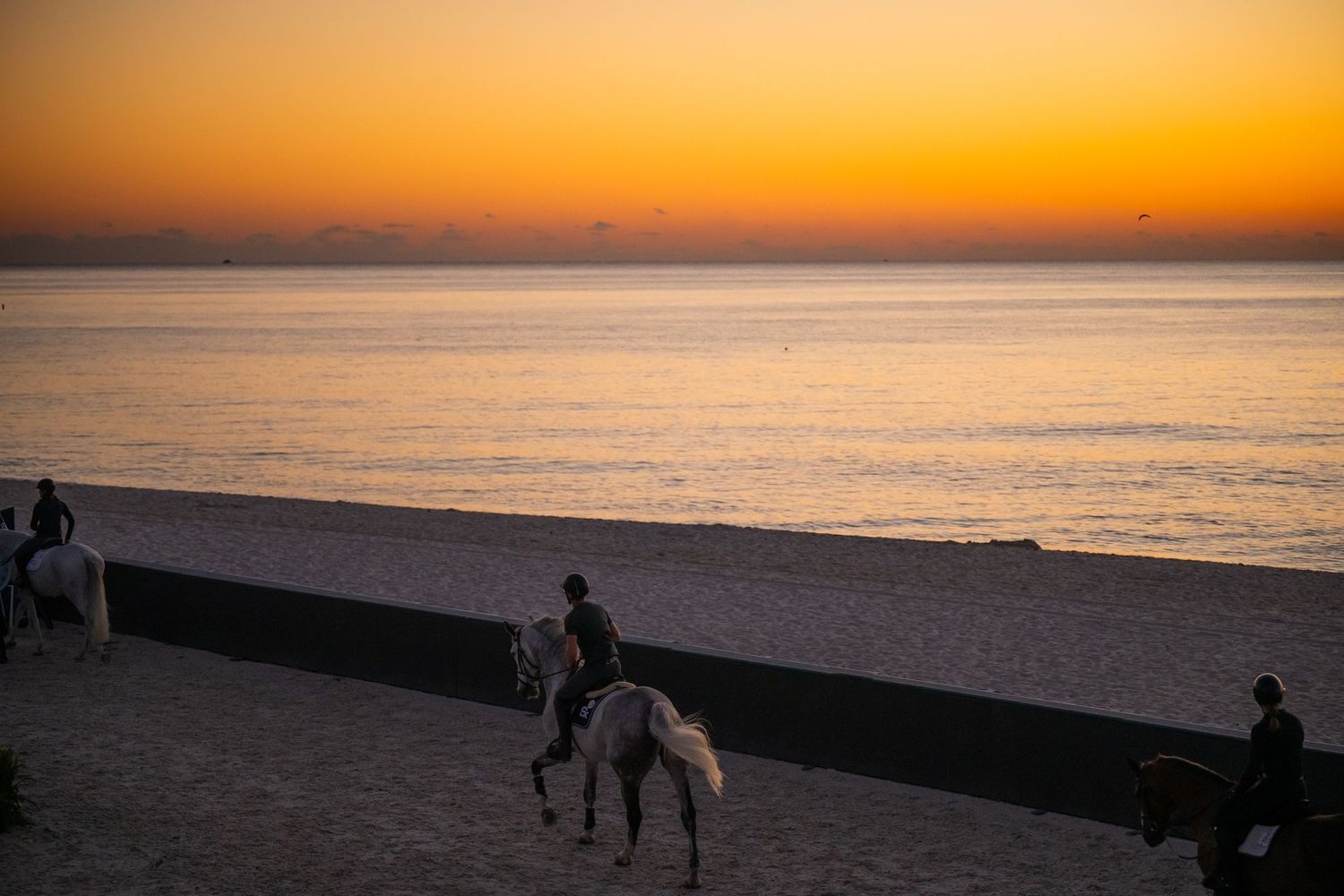 Silhouettes of people and horses walking along a beach at sunset with golden orange sky and ocean waves.