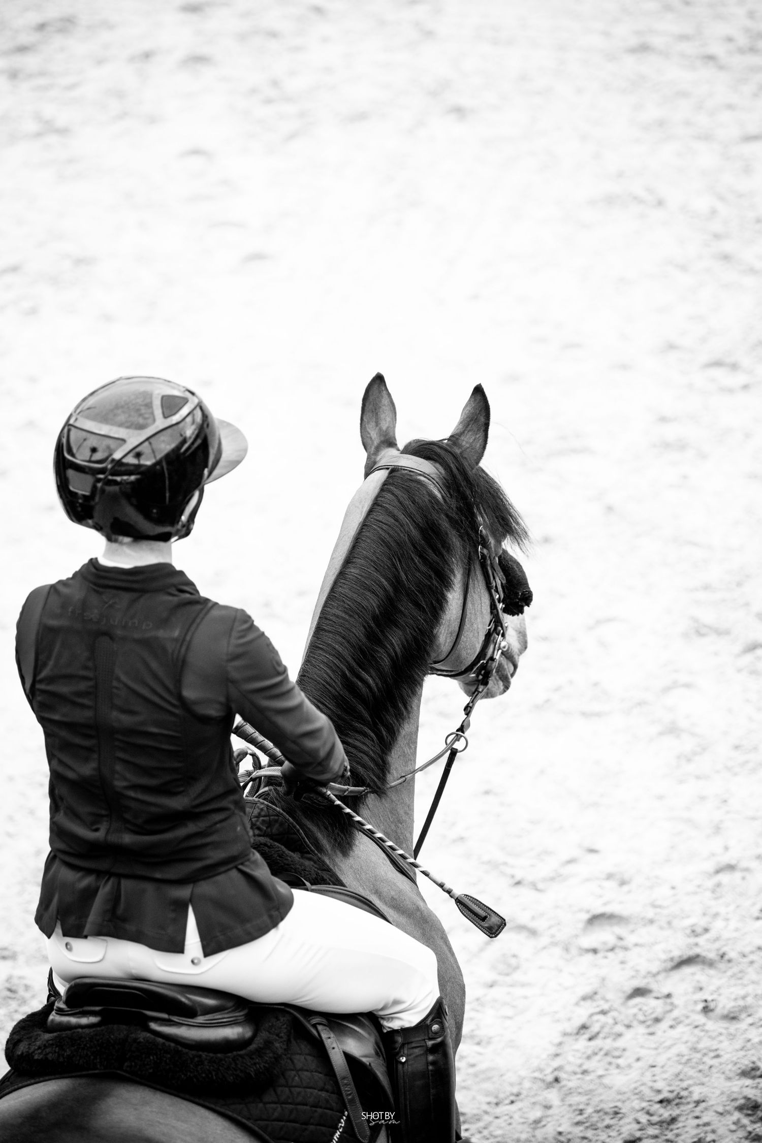 A rider in equestrian gear sits atop a black horse in a sandy arena during competition, captured in black and white.