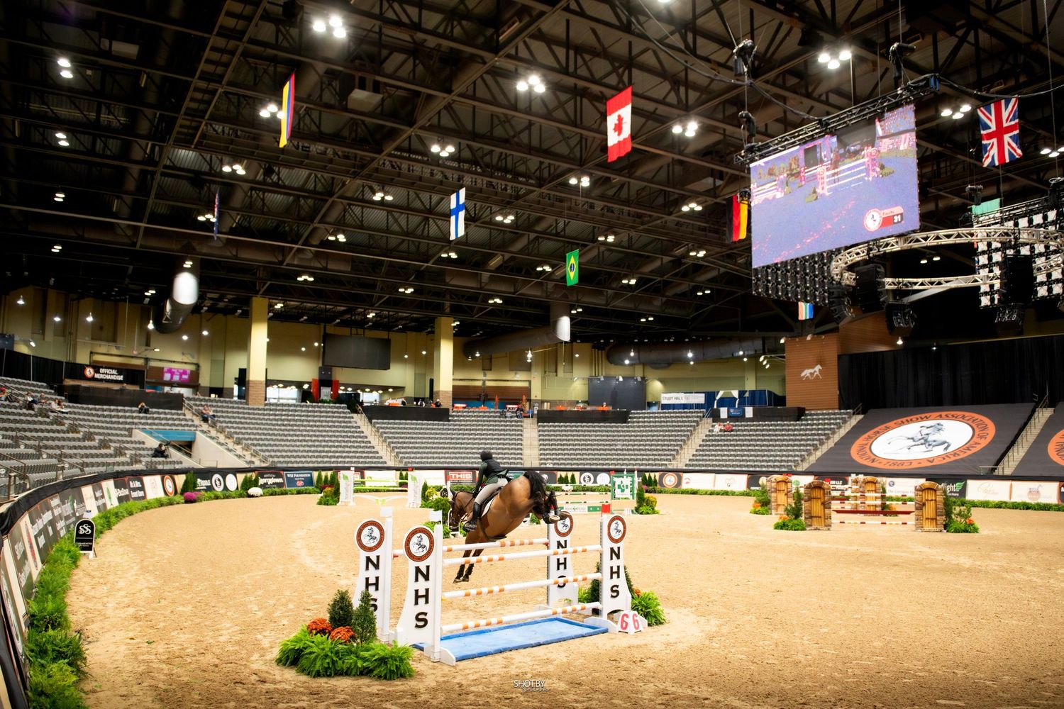 Indoor equestrian arena with multiple international flags hanging from the ceiling during a show jumping competition.