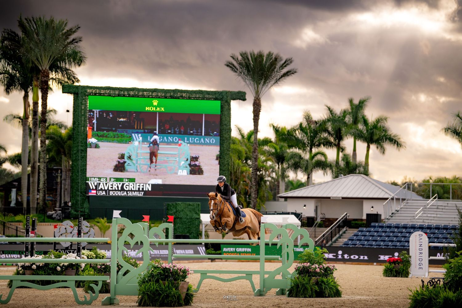 Equestrian show jumping competition at dusk with large video screen and palm trees against dramatic cloudy sky.