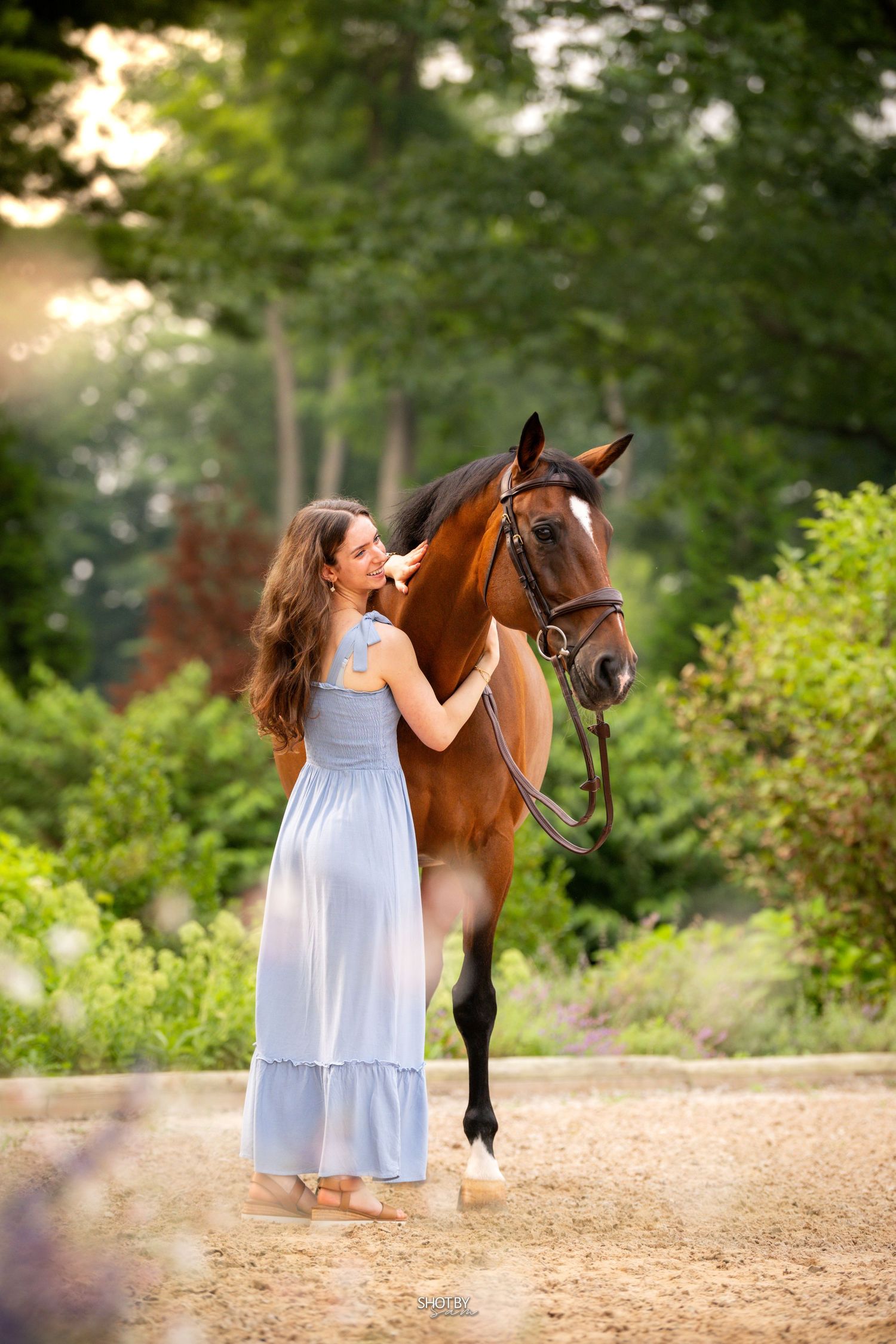 A bay horse stands with someone in a flowing blue dress on a dirt path surrounded by lush greenery.
