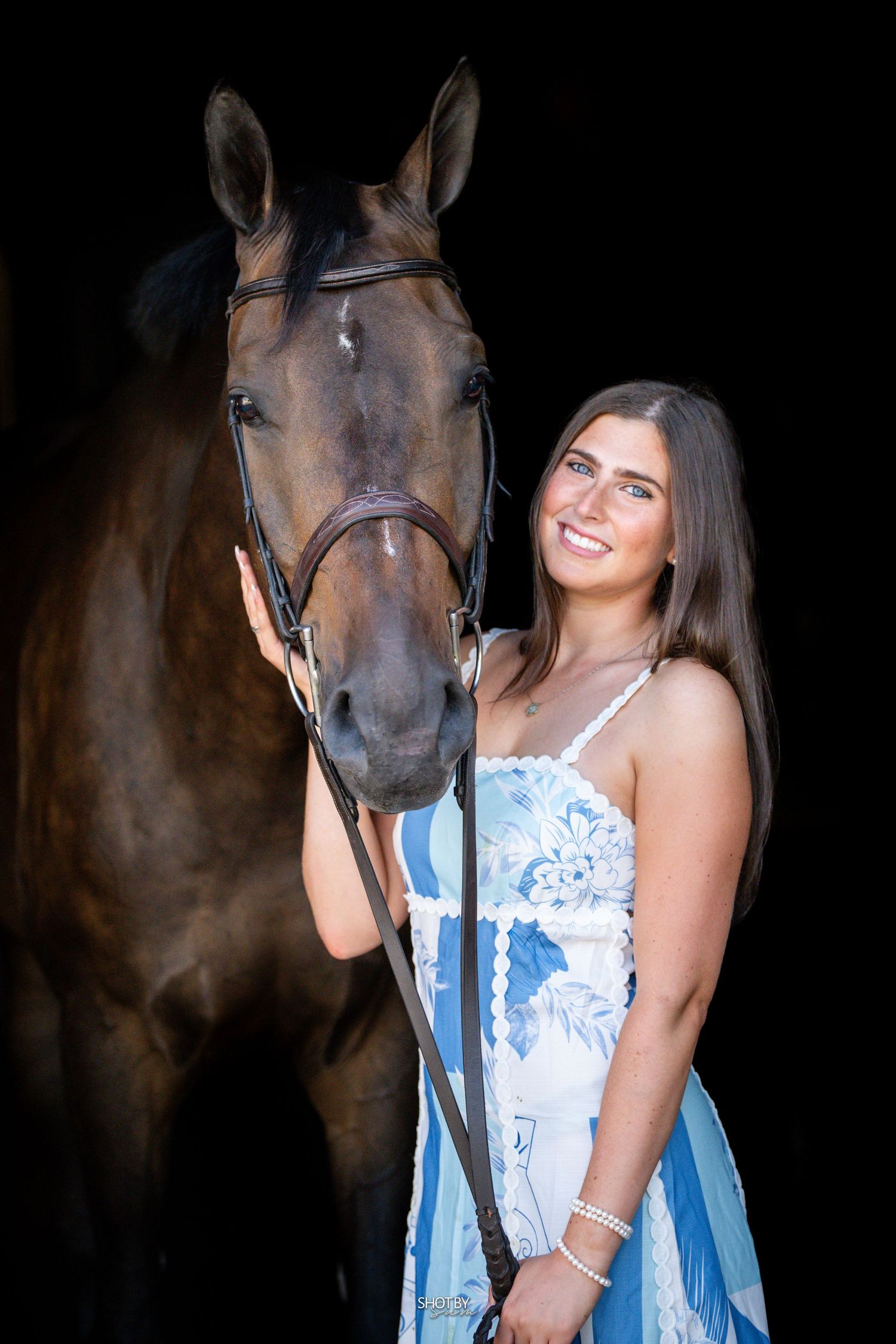 A bay horse with bridle poses against dark background while being shown affection.