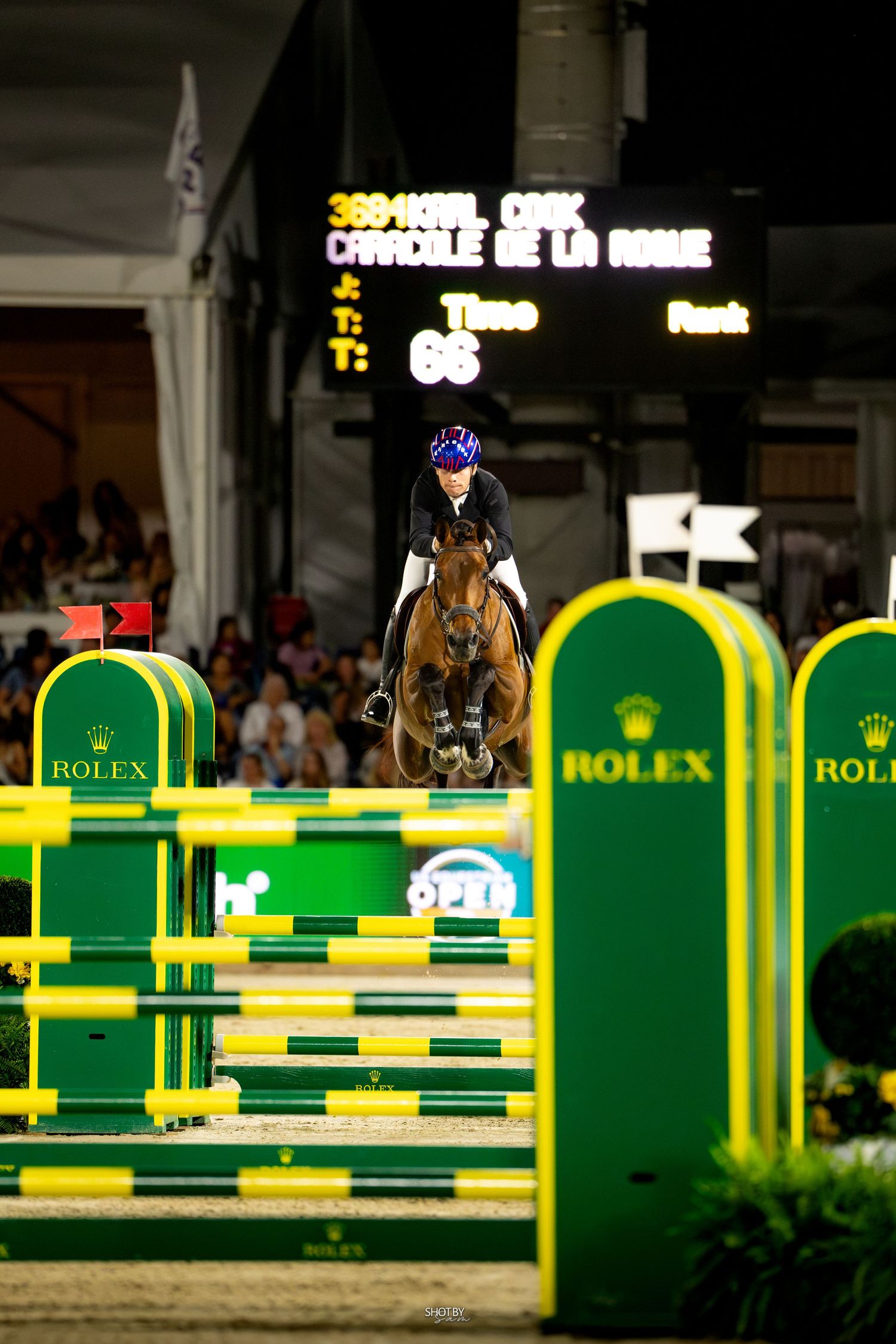 Equestrian rider jumps over yellow and green obstacles during a Rolex sponsored show jumping competition.