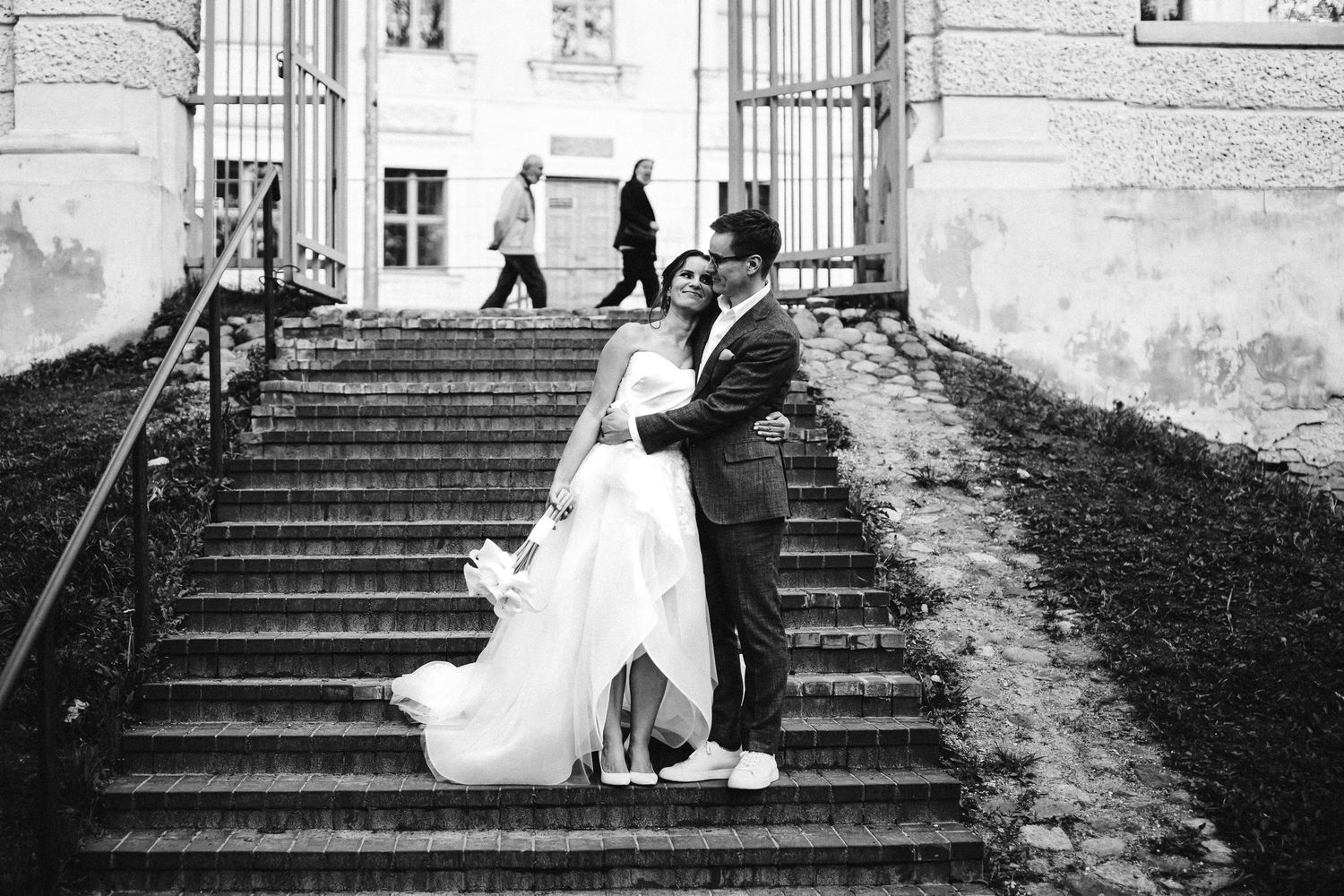 Jaunavedžiai pozuoja nuotraukai ant pastato laiptų, šypsosi ir laiko vienas kitą už rankų.
