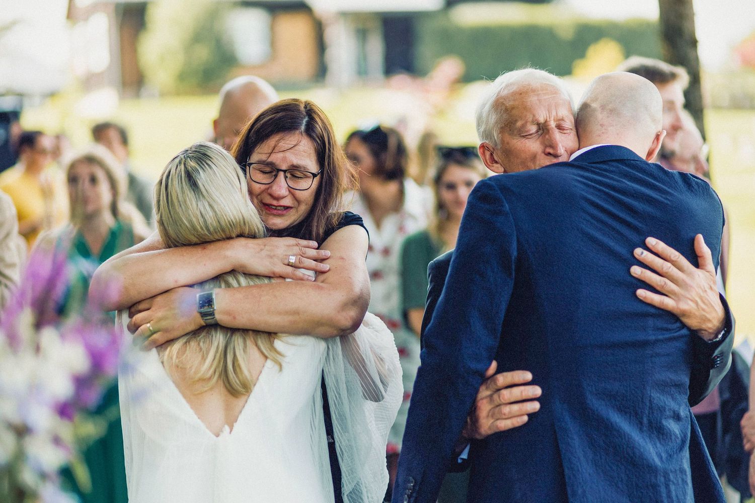 Jaunavedžiai ir jų tėvai apkabina vienas kitą savo vestuvių ceremonijoje, šypsodamiesi ir džiaugdamiesi.