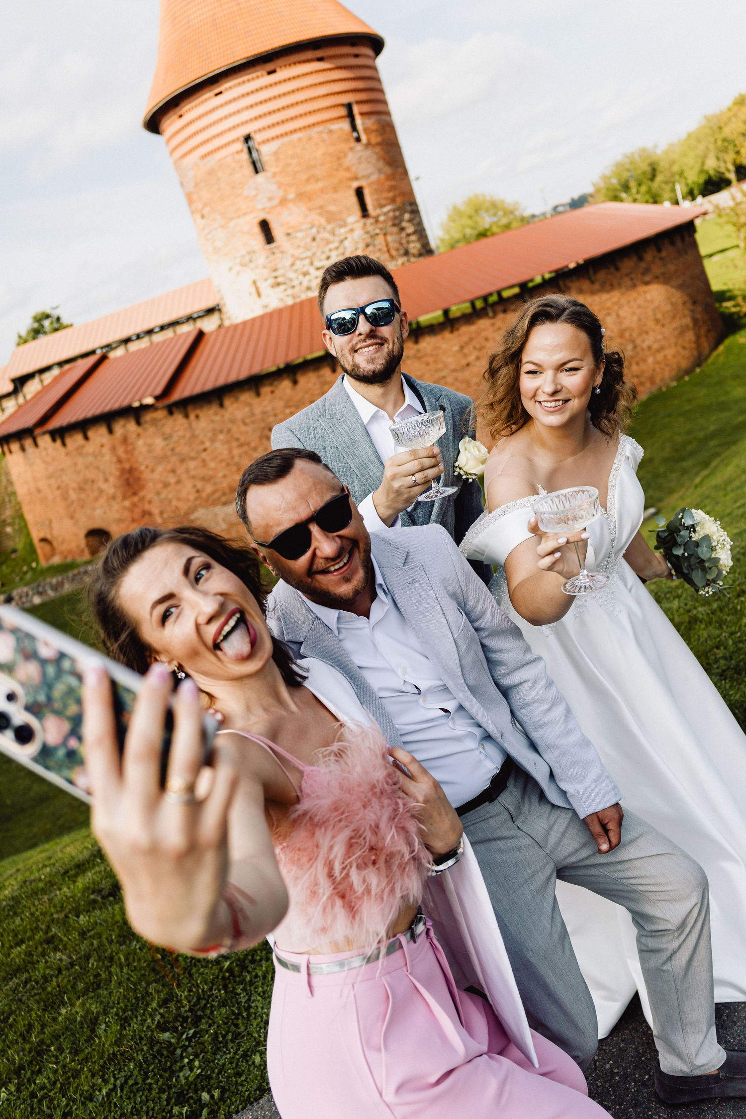 Grupė vestuviš svečiudaro selfie prieš Kauno pilį. Visi šypsosi ir džiaugiasi akimirka.
