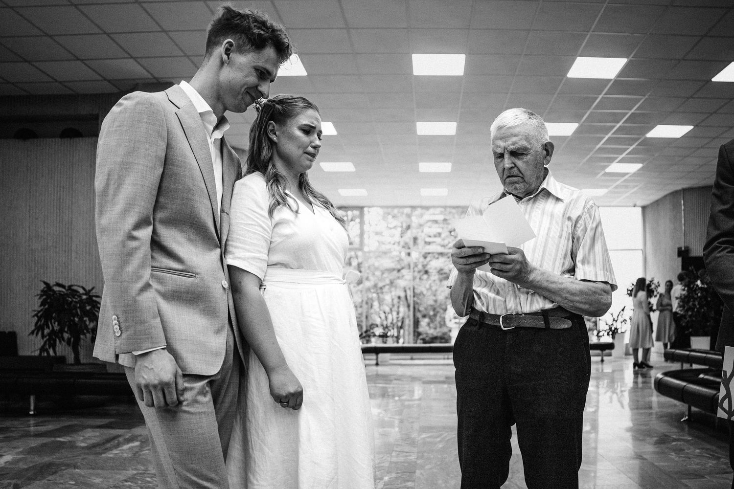 Vyras ir moteris stovi prabangiame fojė Vilniaus santuokų rūmuose,  su seneliu, aplinka elegantiška ir rafinuota.