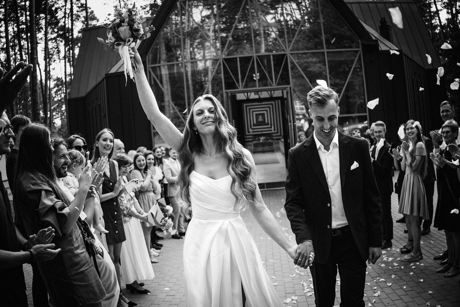 Jaunavedžiai, apsirengę elegantiškais drabužiais, laimingai žengia per prašmatnų vestuvių taką, svečiai ploja