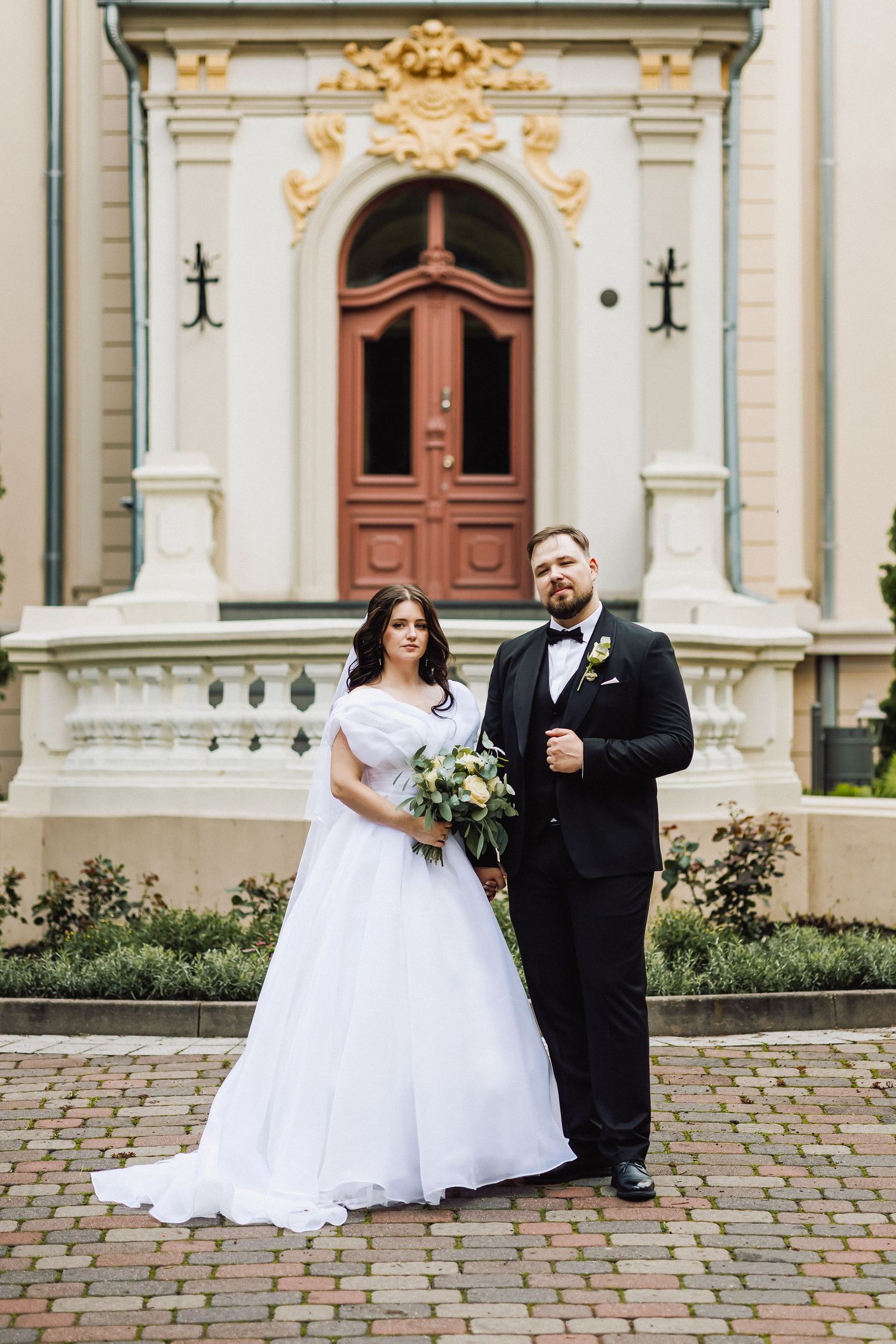 Jaunavedžiai pozuoja nuotraukai prieš bažnyčią, šypsodamiesi ir laikydami vienas kitą už rankų.