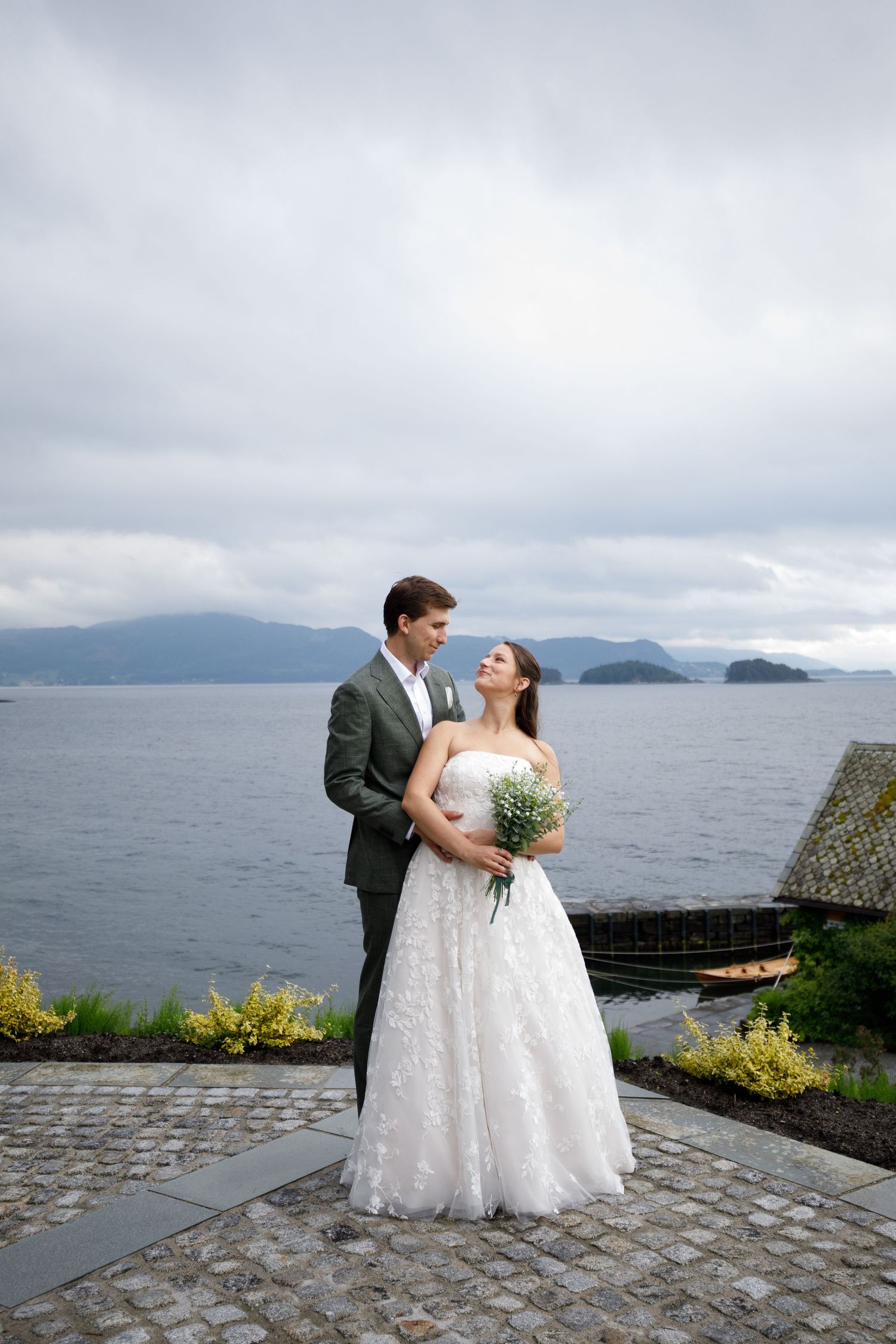 Bride and groom embrace by a scenic lakeside with mountains in the background on their wedding day.