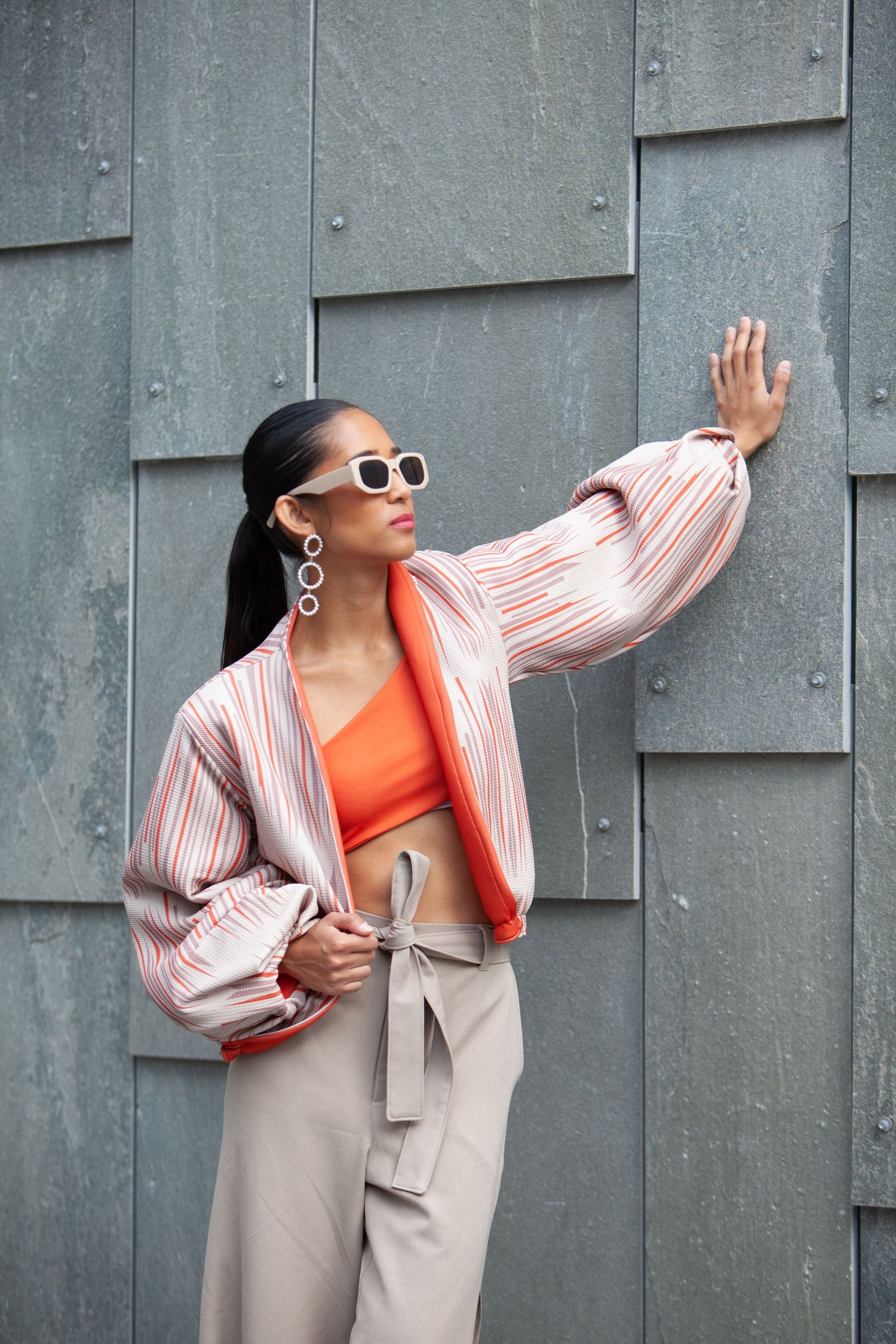 Stylish woman in orange top and striped blazer poses against gray wall in fashion photoshoot series.