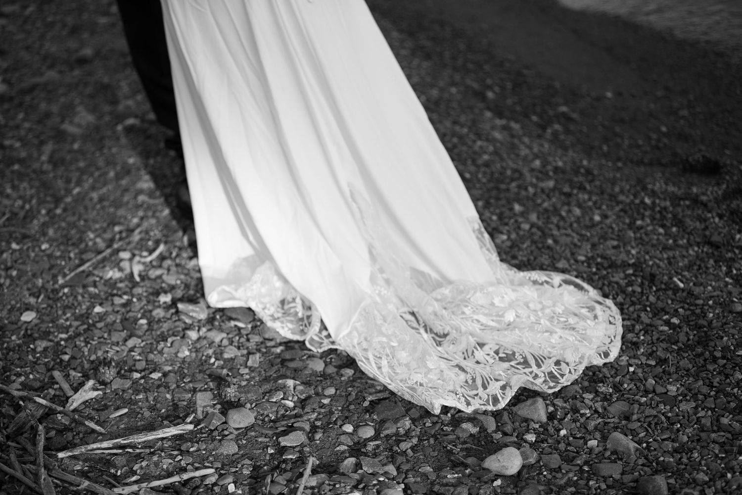 A flowing white wedding dress train with delicate lace details trails along a dark stone path.