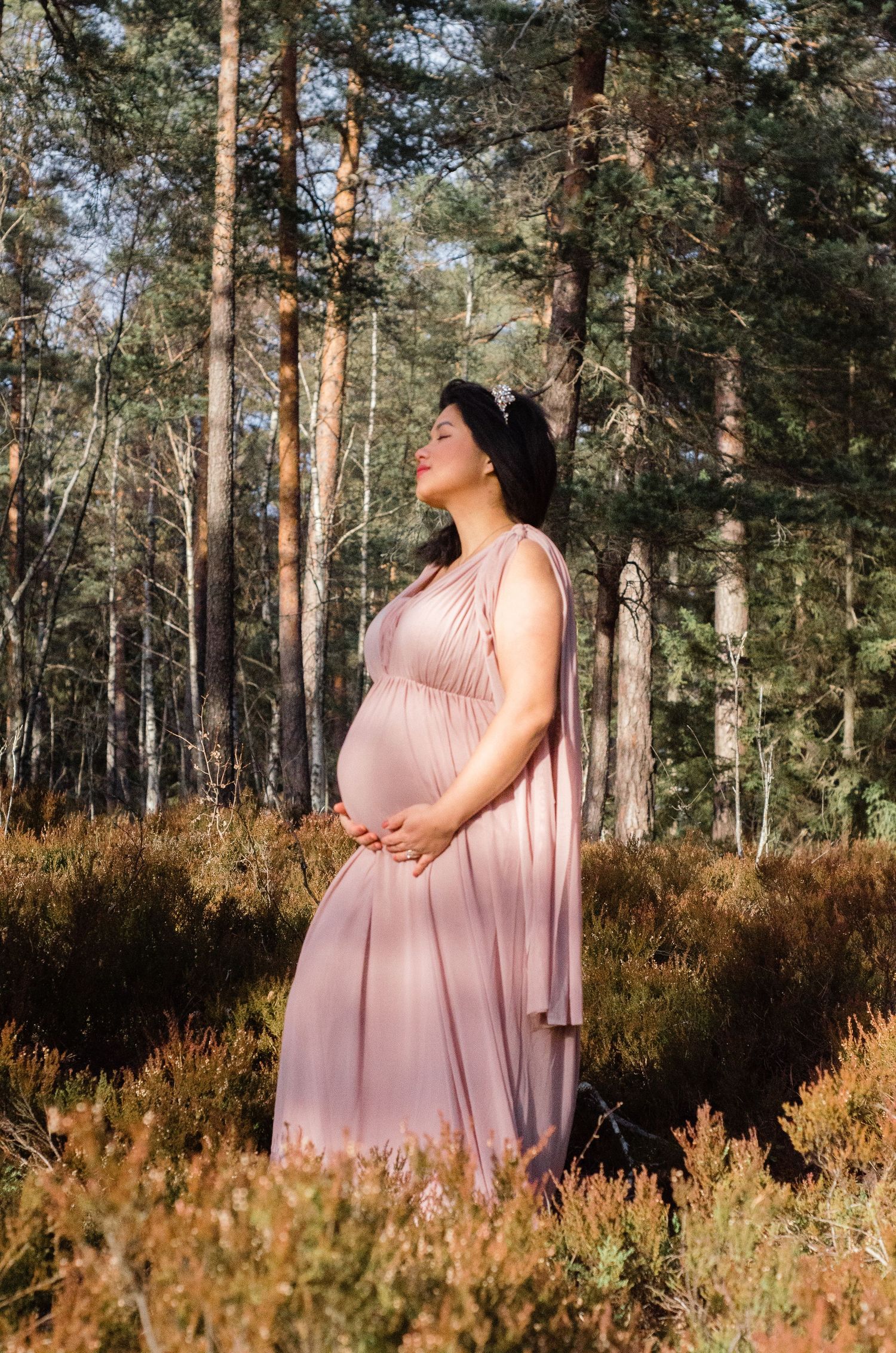 Maternity photoshoot in a forest with pink dress and natural lighting among pine trees and heather.
