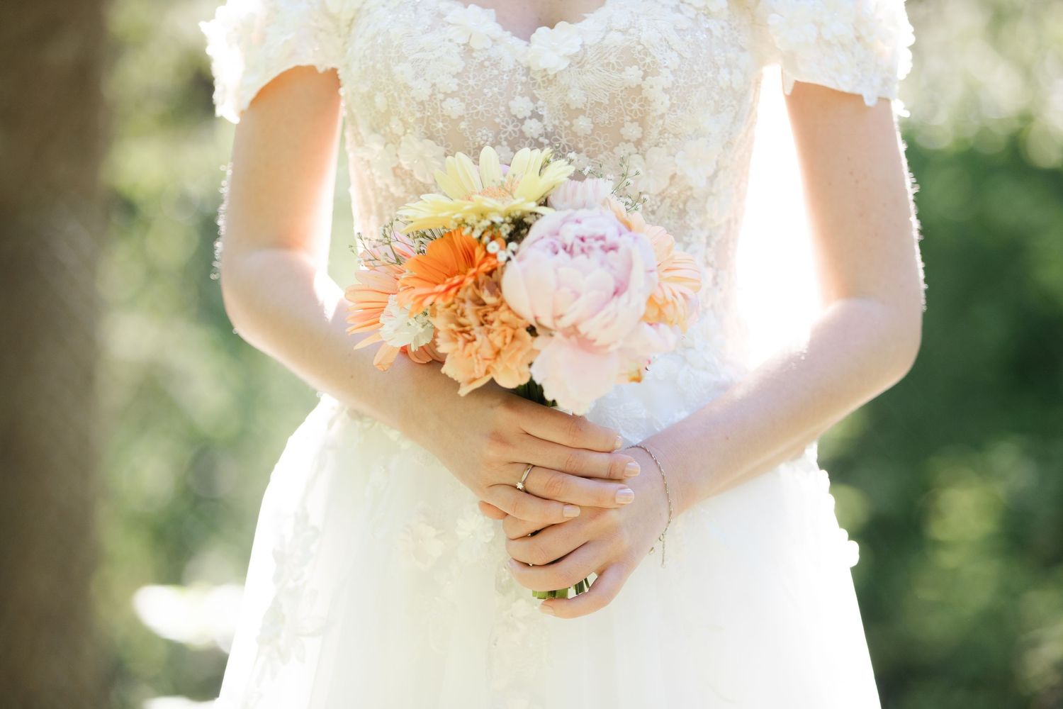 A delicate bouquet of pink and white flowers is held against a white lace wedding dress in natural sunlight.