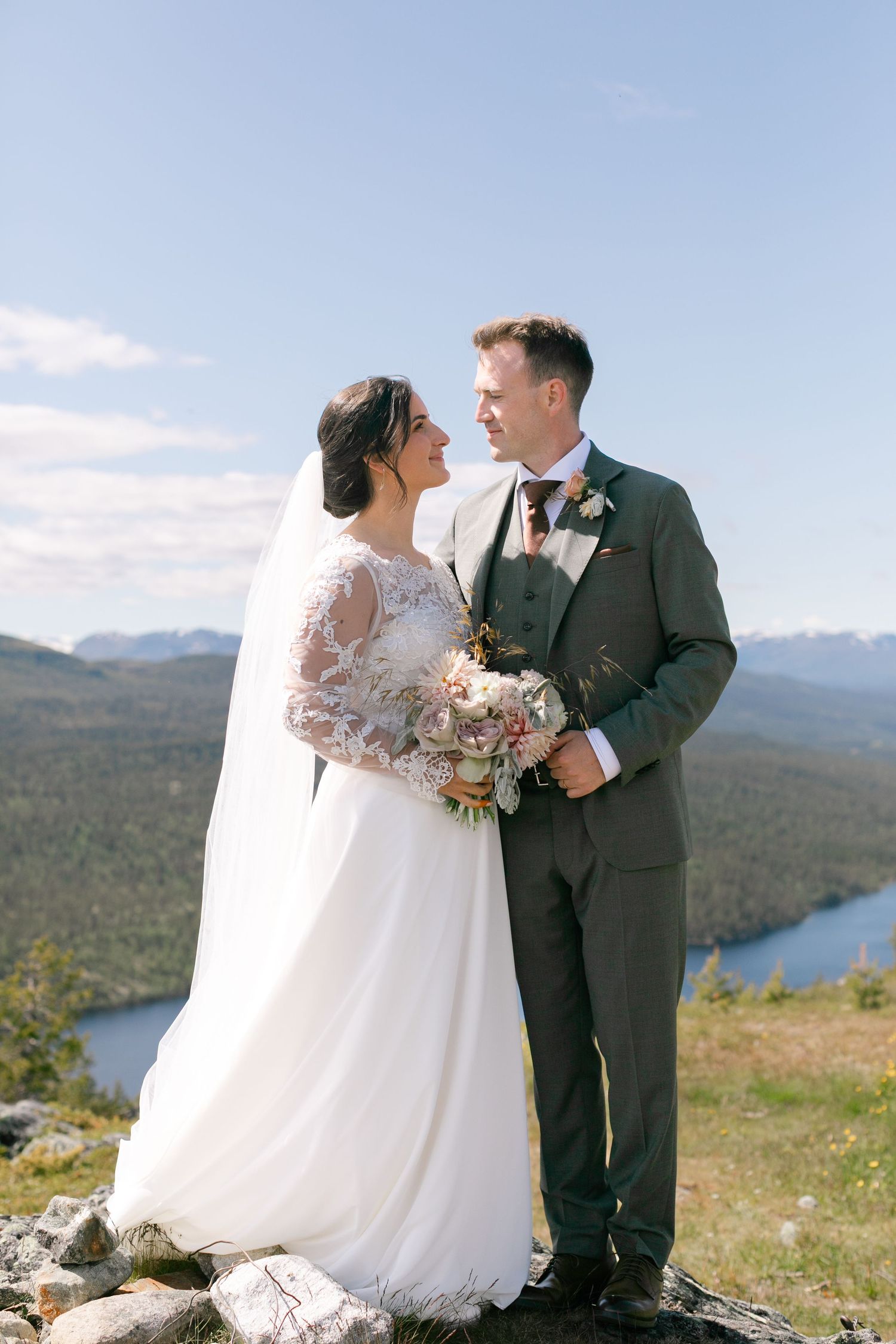 Wedding couple poses on mountaintop with scenic lake and mountain views in background.