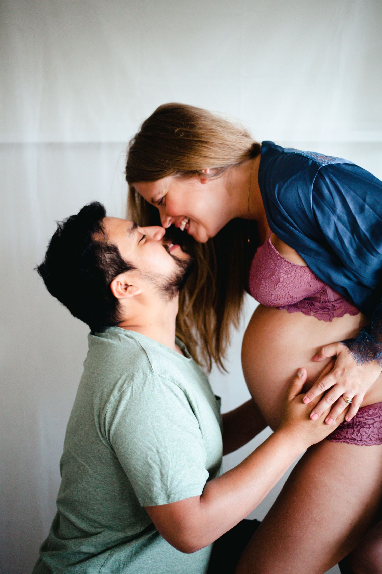 Intimate maternity photography session with couple in casual attire against white backdrop.