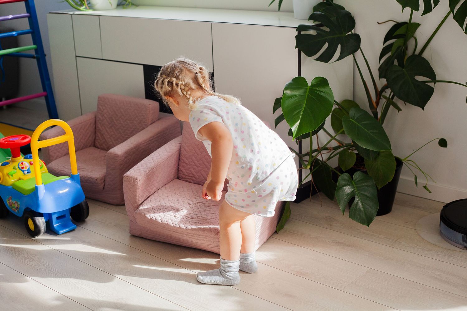 A small child in white clothing exploring a pink chair next to indoor plants.