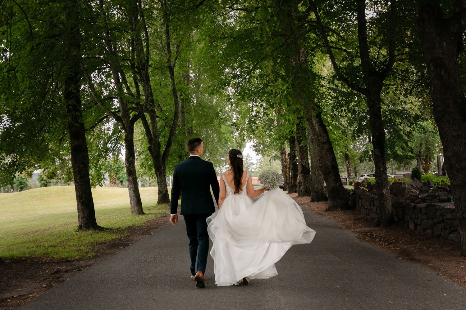 Tree-lined pathway in a park with wedding couple walking through the natural archway.