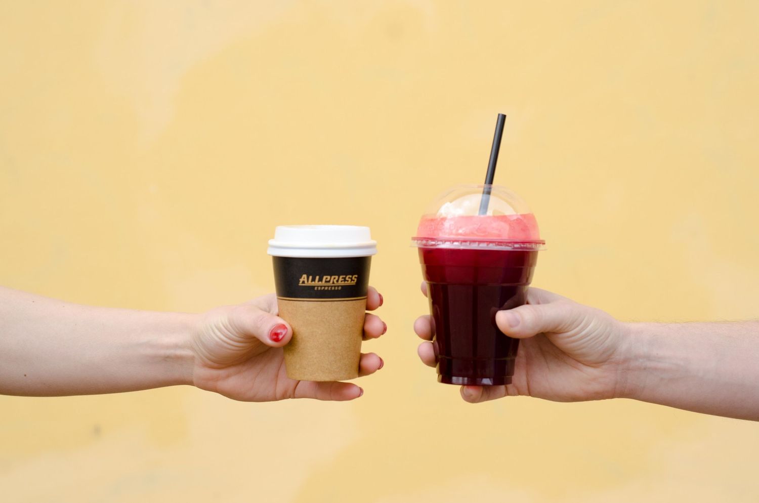 Two hands reach out to exchange colorful beverages in plastic cups against a yellow background.