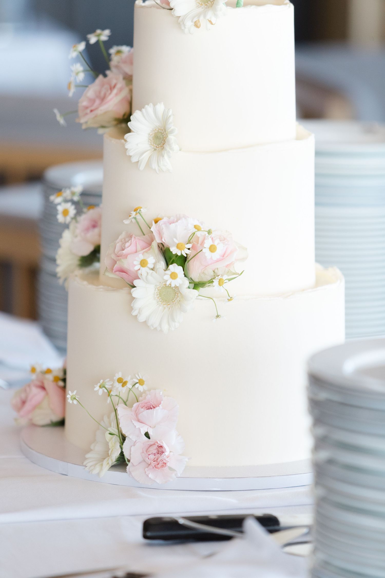 Elegant three-tier white wedding cake decorated with fresh pink roses, white daisies and delicate baby's breath flowers.
