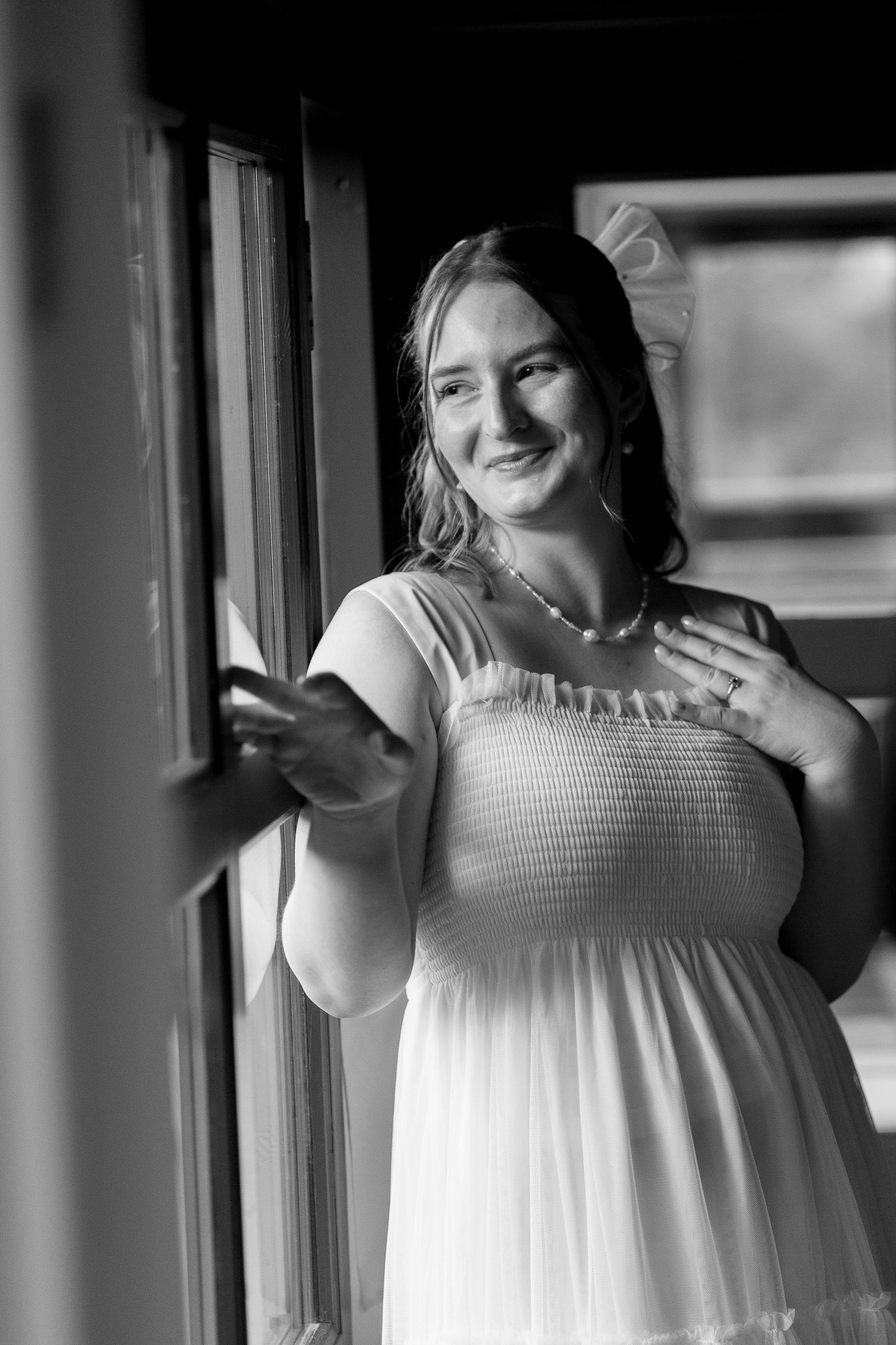 A person in a flowing white dress poses joyfully near a window in black and white photography.