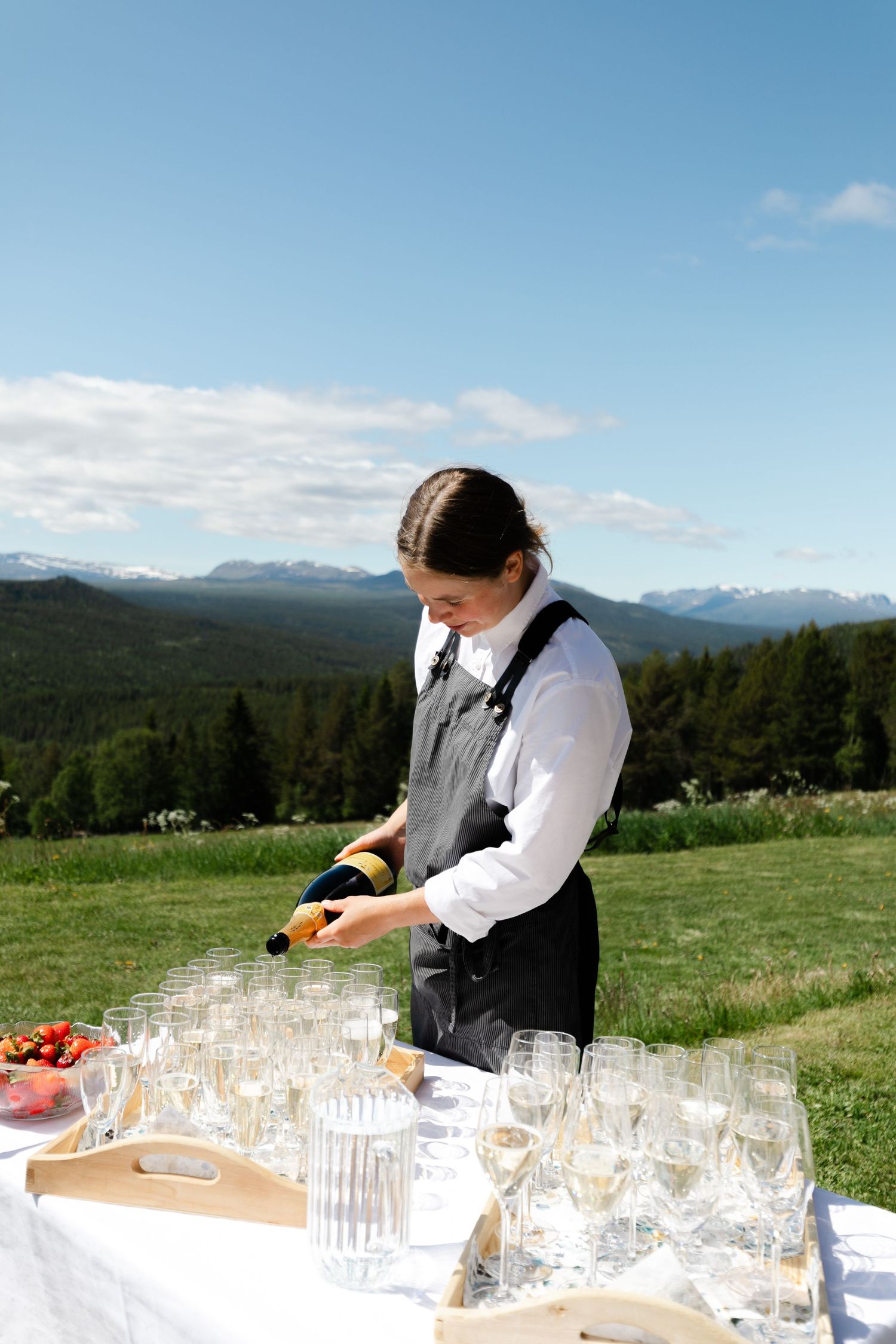 Catering staff preparing drinks on tables outdoors with mountain landscape in background.
