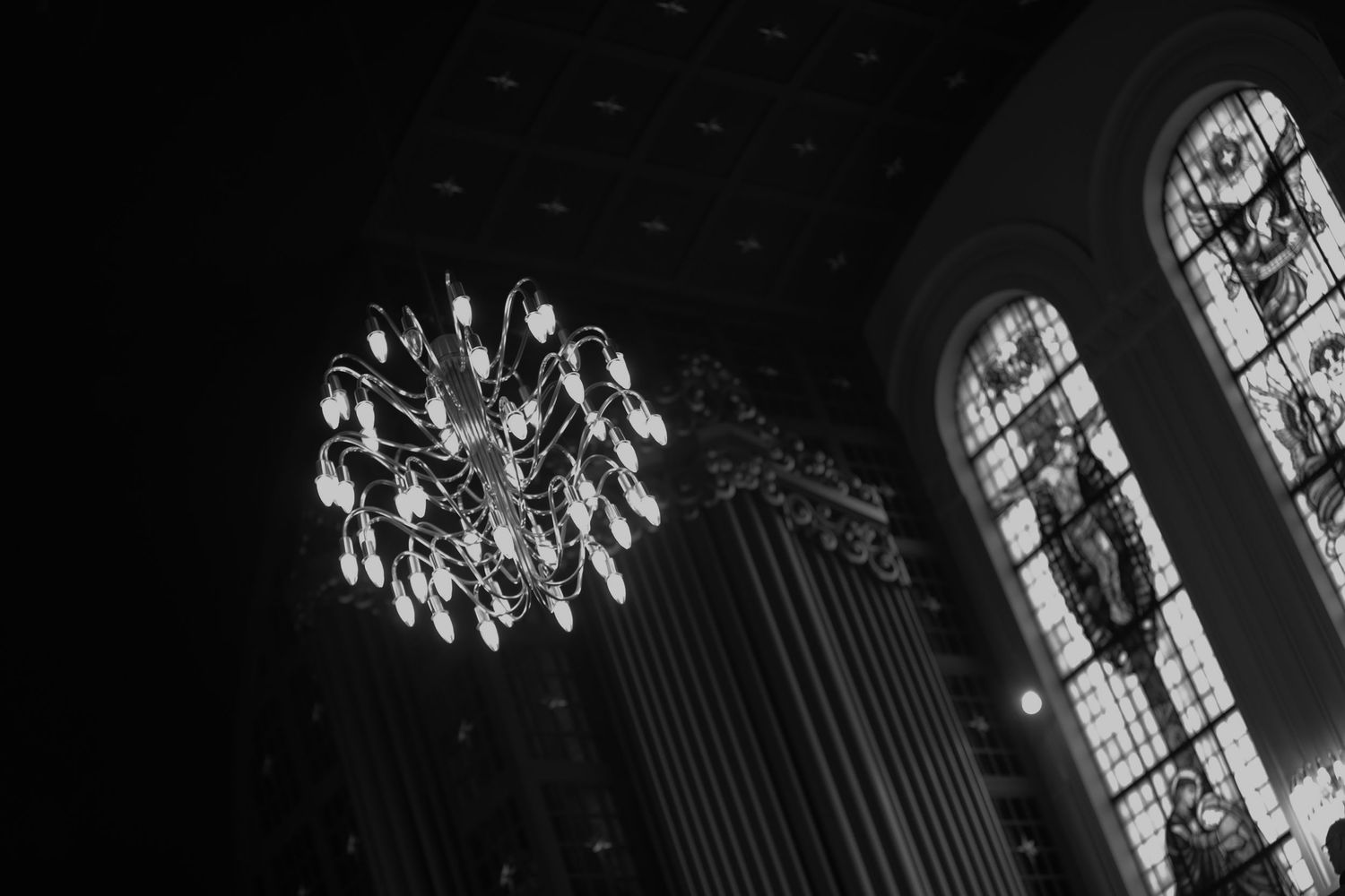 Black and white photo of an elegant chandelier and stained glass windows inside a grand church.