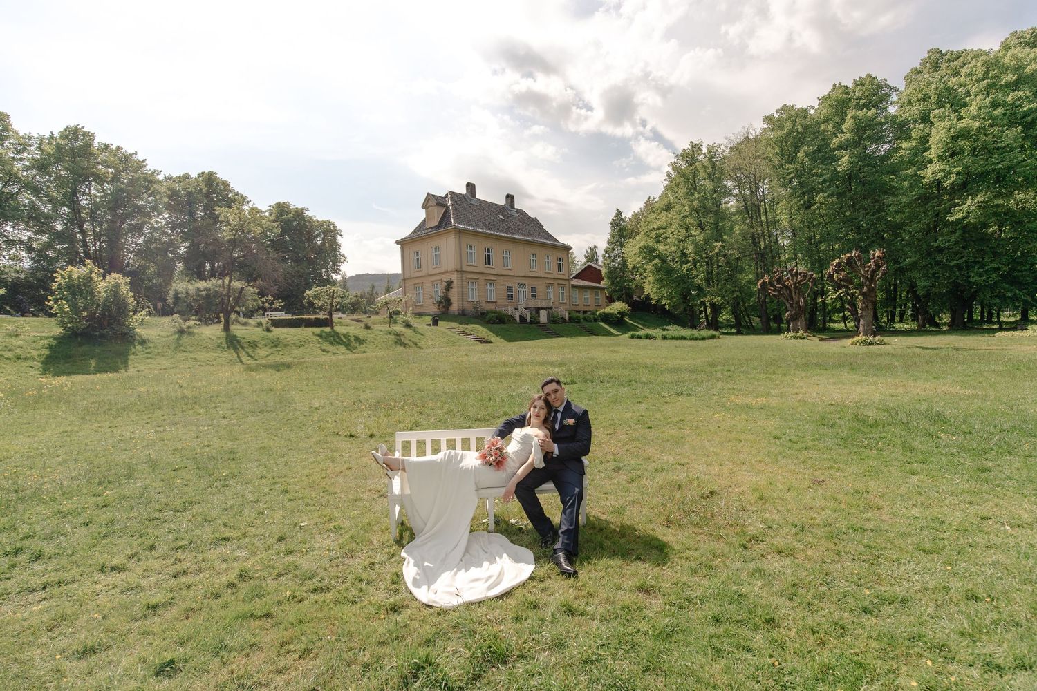 Bride and groom pose romantically on a lush green lawn with a historic stone manor and tall trees in the background.
